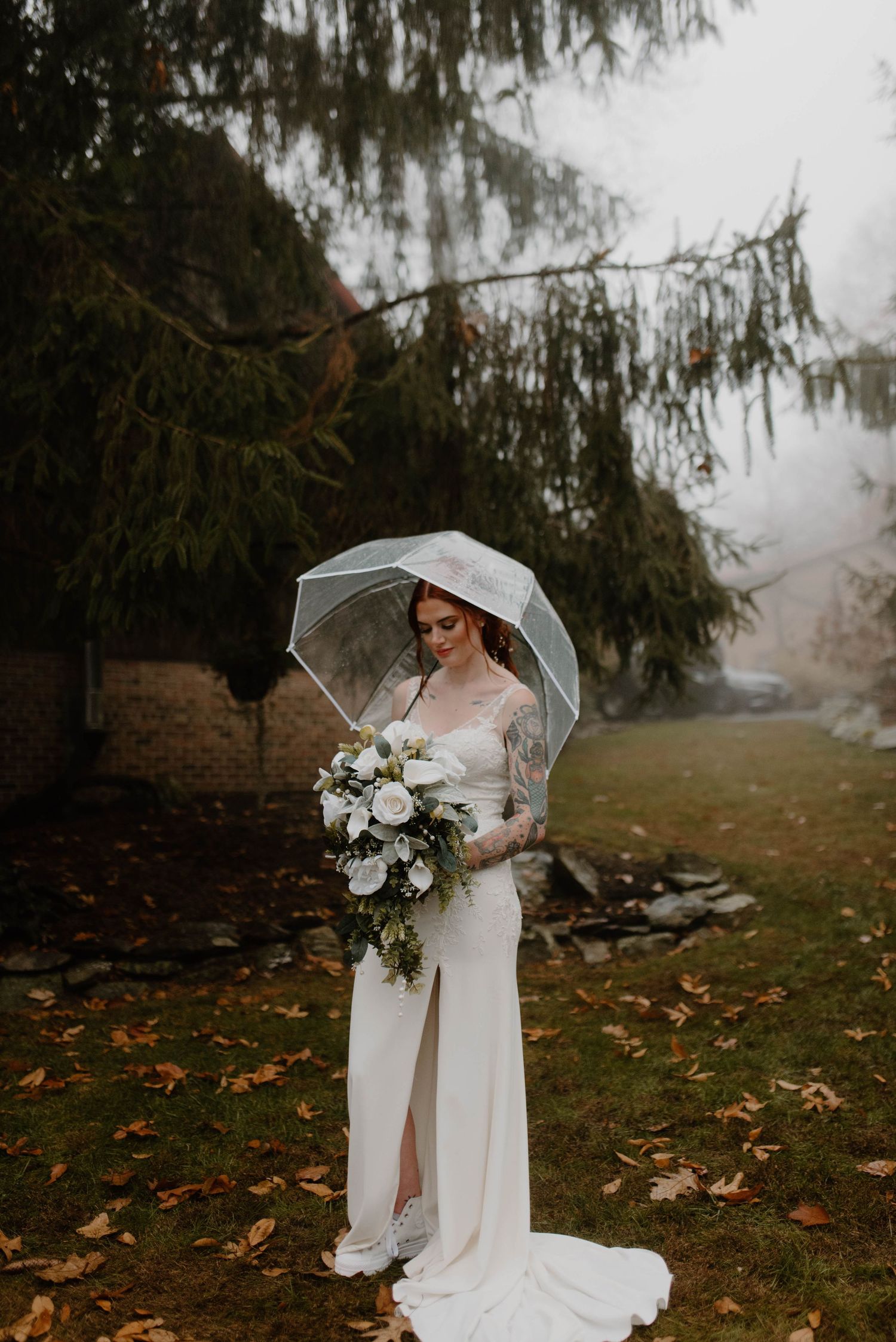 A bride in a sleek white dress holds a bouquet under a clear umbrella on a misty autumn day.