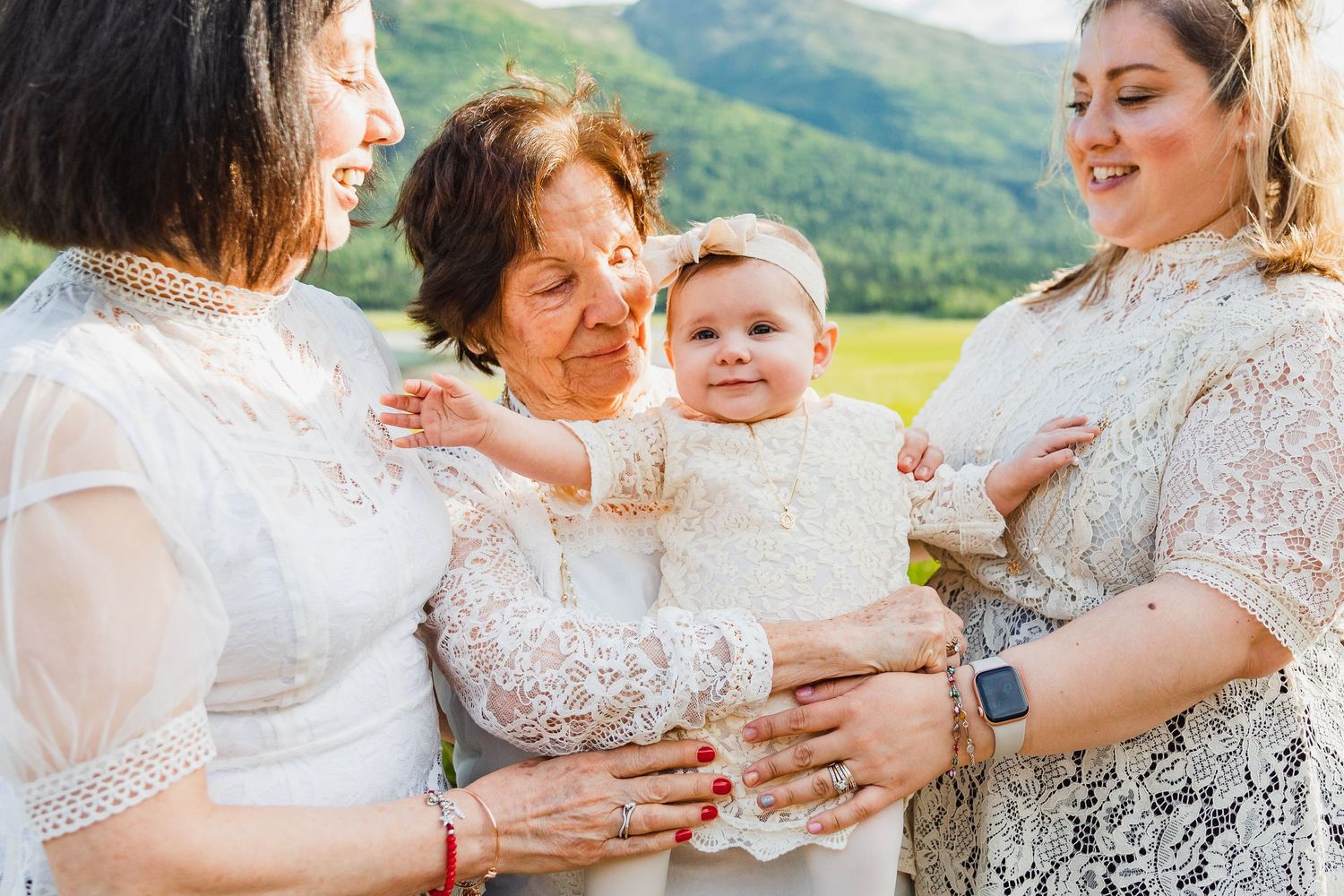 Three generations share tender moment with baby in white lace against mountain backdrop.