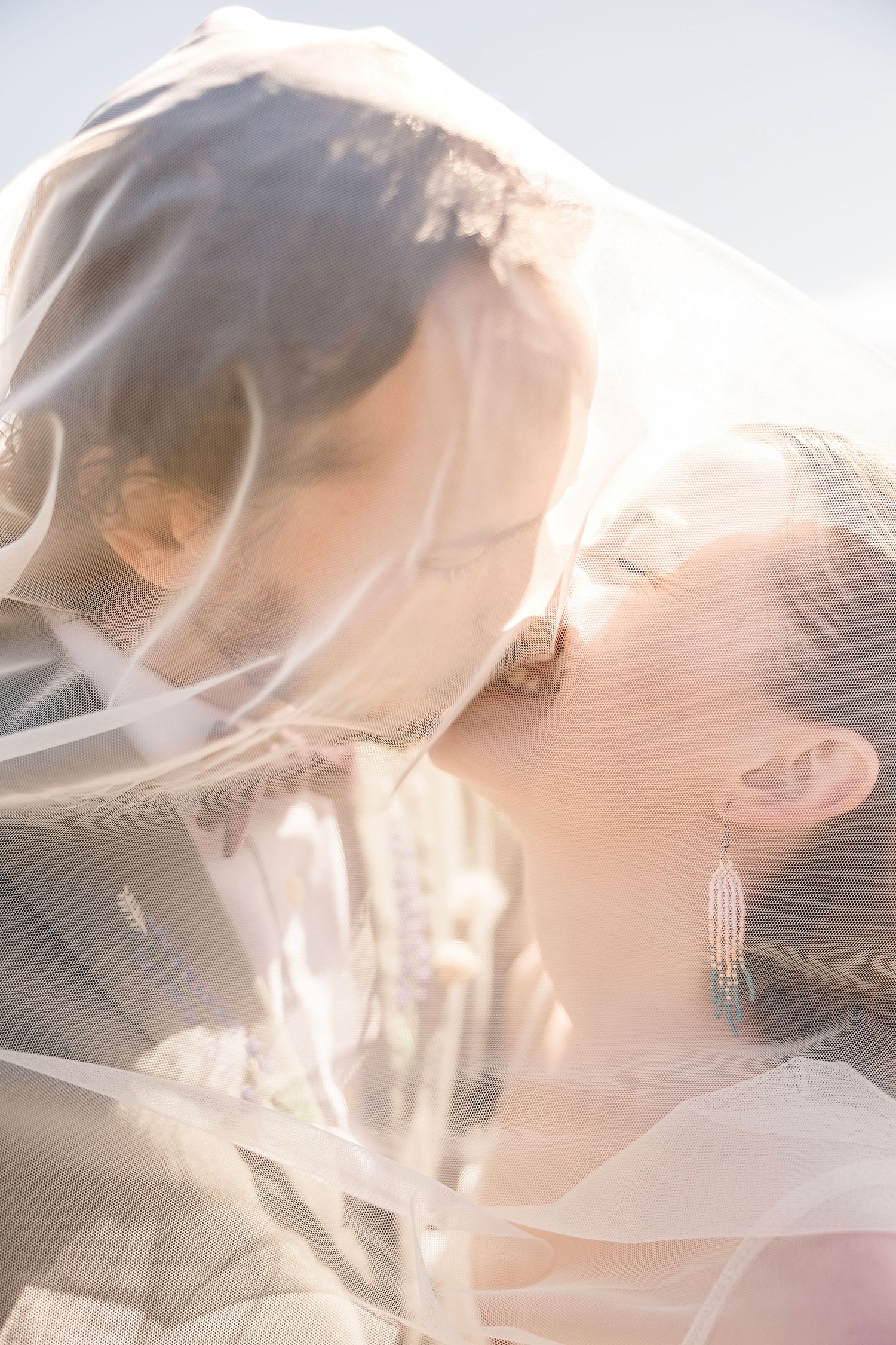 Ethereal close-up shot of a bride under flowing white veil with soft sunlight creating a dreamy atmosphere.