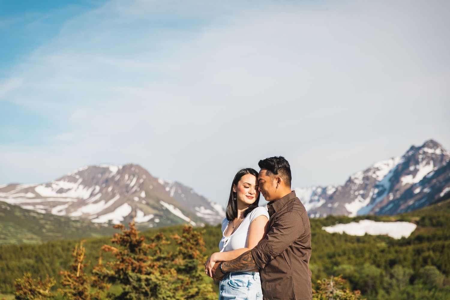 Couple embraces against backdrop of snow-capped mountains and wildflowers in an alpine meadow at sunset.