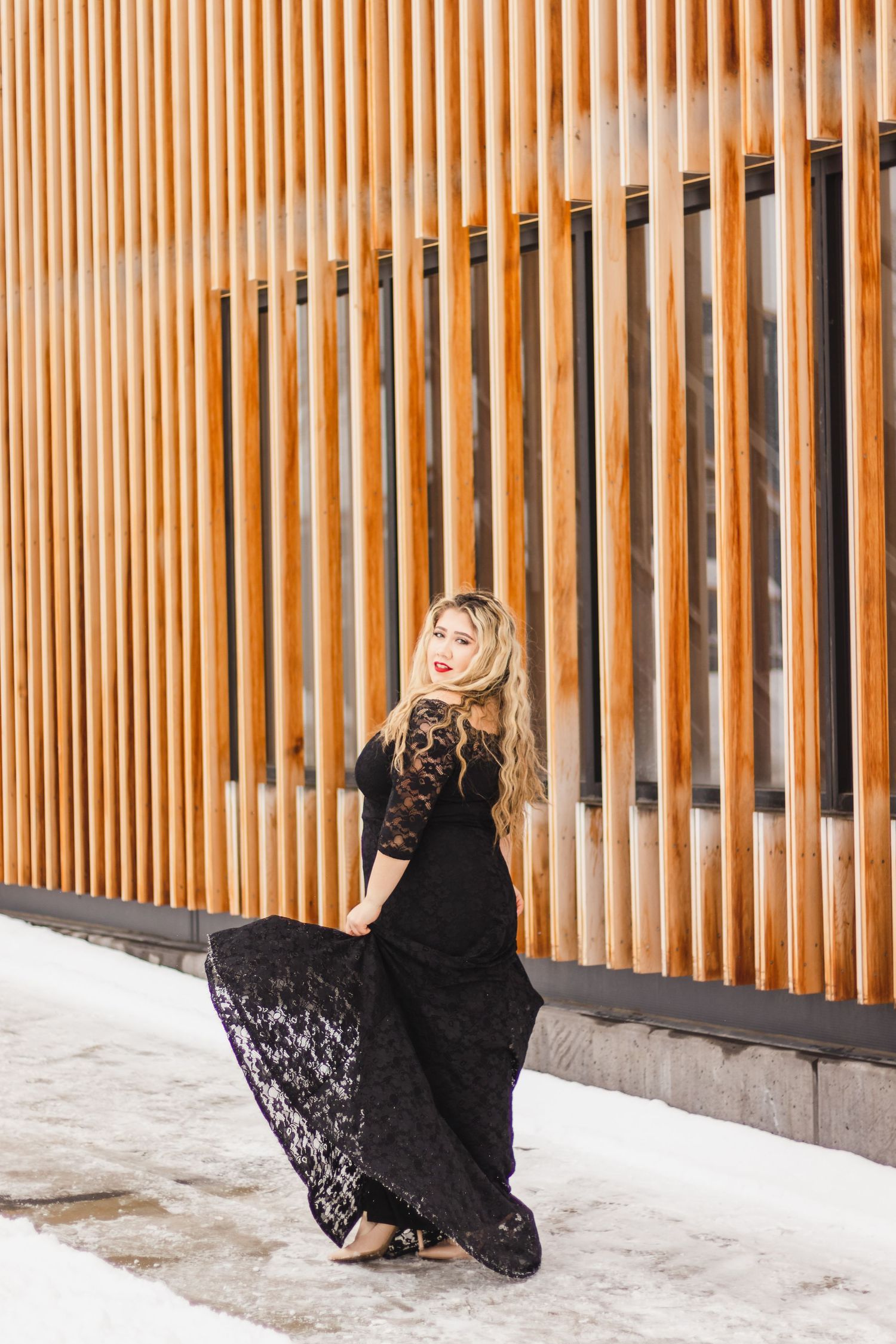 Person in black lace dress twirling near modern wooden slatted wall in winter.