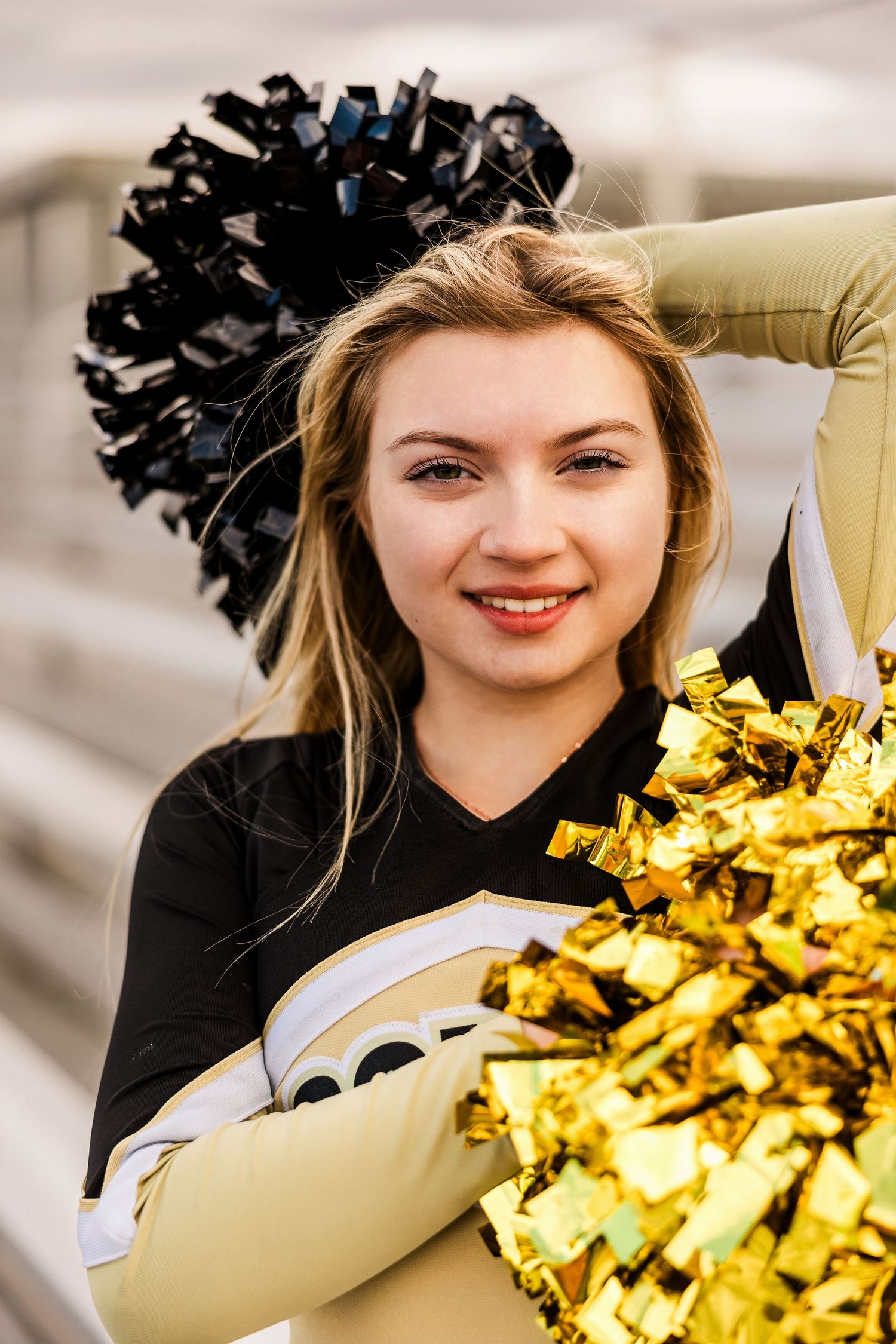 A cheerleader in black and white uniform holds gold pom poms while smiling during a photo session.