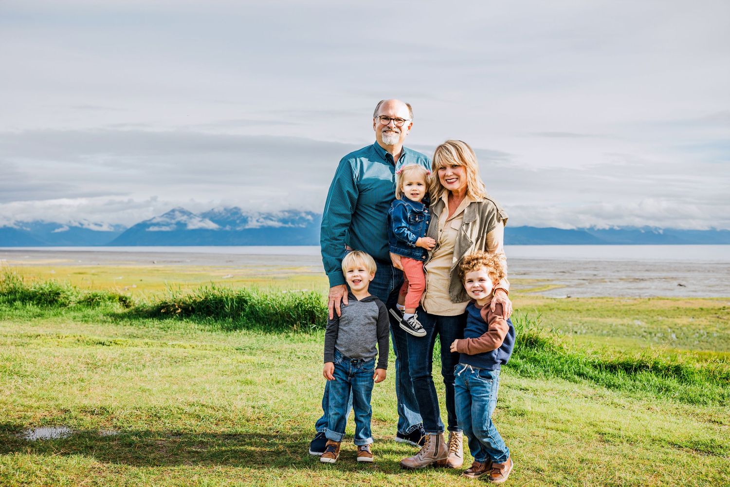A group stands together on a scenic grassy hillside overlooking a mountain range and bay on a sunny day.