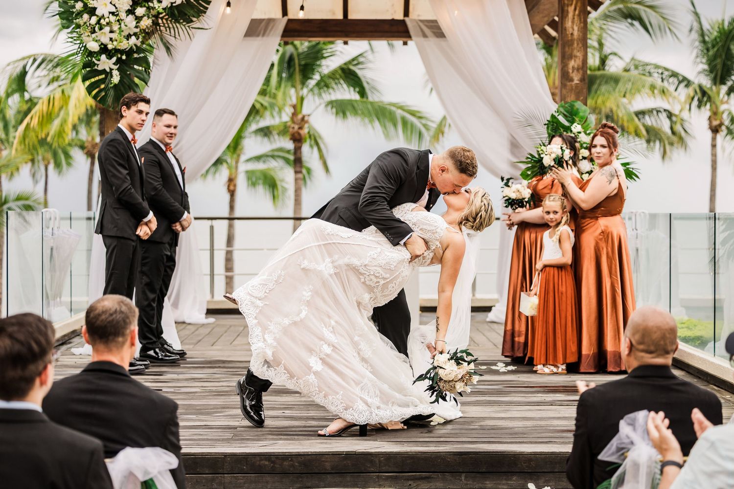 Wedding party celebrates on a beachfront terrace with palm trees and flowing white drapery in the background.
