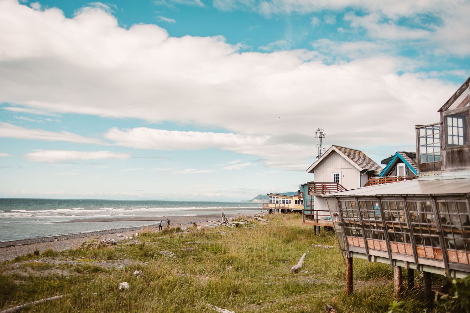 Wooden beachfront homes and boardwalk along a sandy coastline under blue skies.