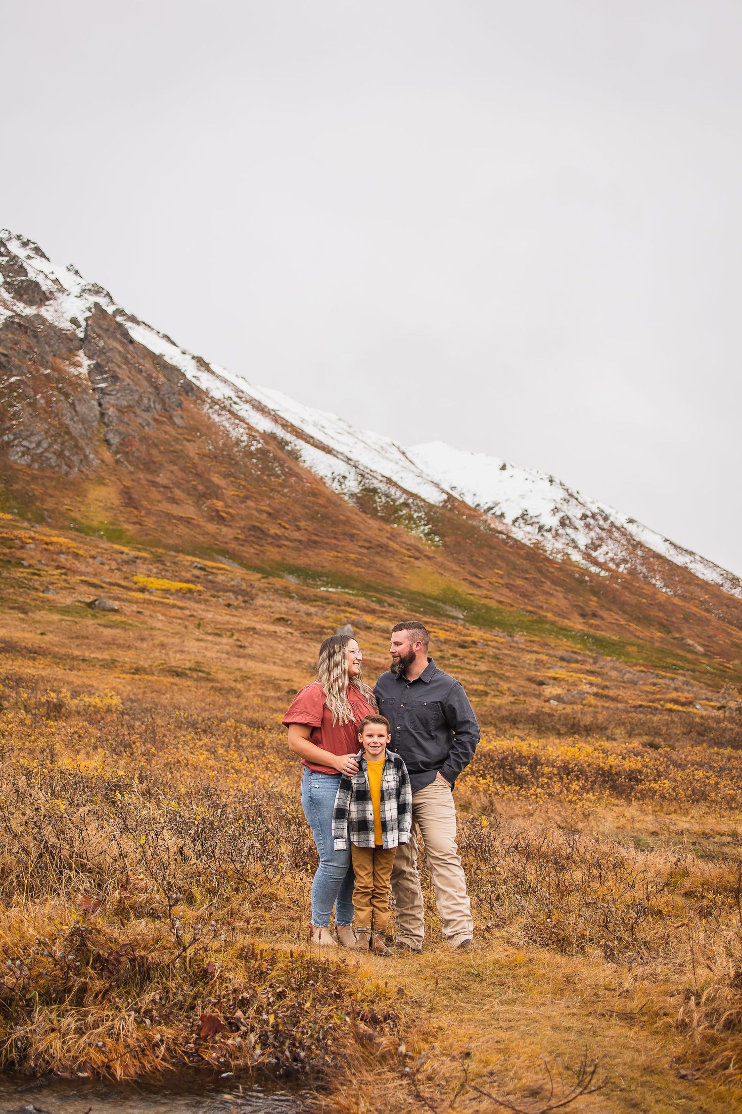 Couple standing together on autumn mountain trail with snow-capped peaks in background.