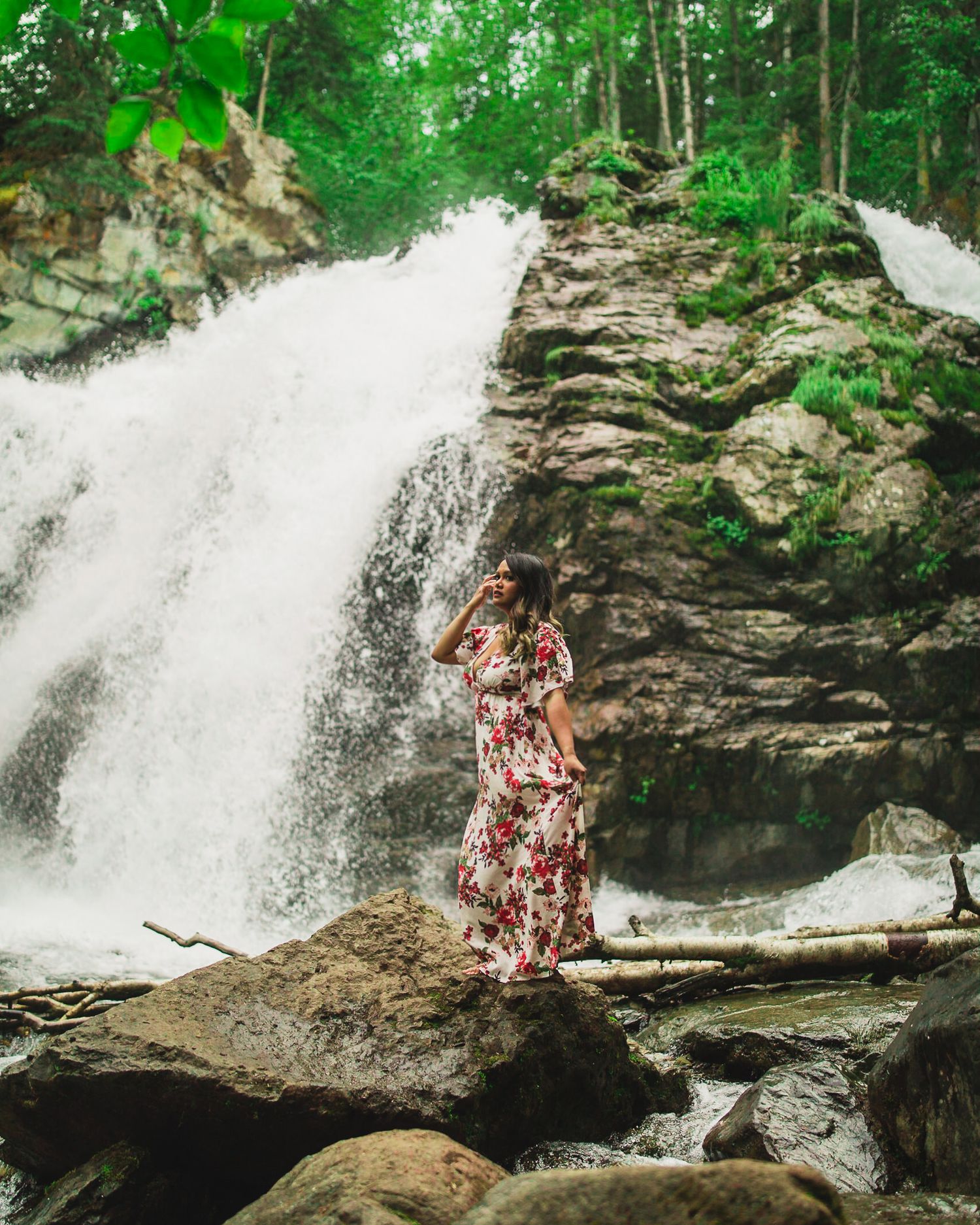 A figure in a floral dress poses near a cascading waterfall surrounded by lush green forest.