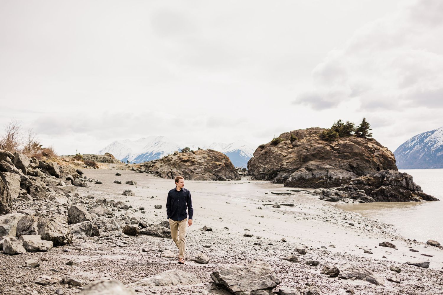 Person walking along rocky coastal landscape with mountains in the distance.