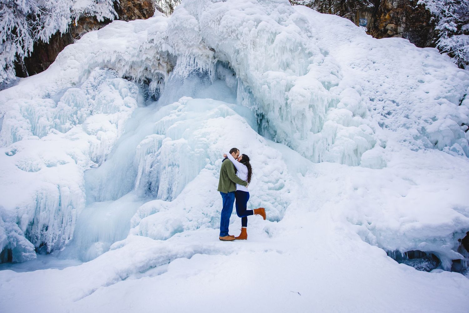 Two figures stand in front of a massive frozen waterfall with blue and white ice formations during winter.