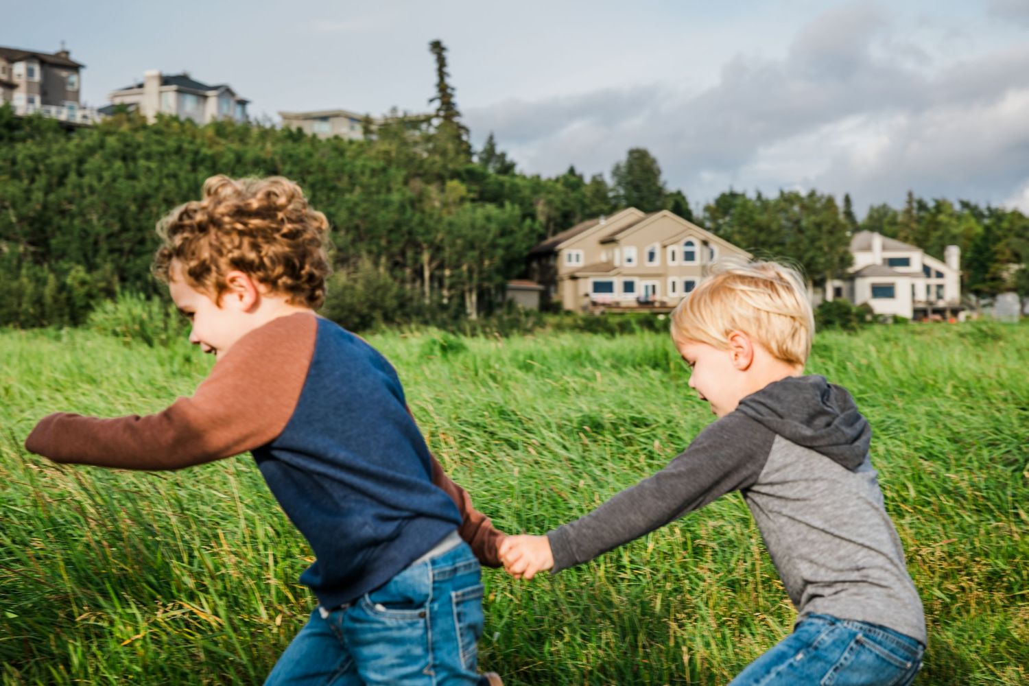 Two kids playing and running through a grassy field with houses visible in the background.
