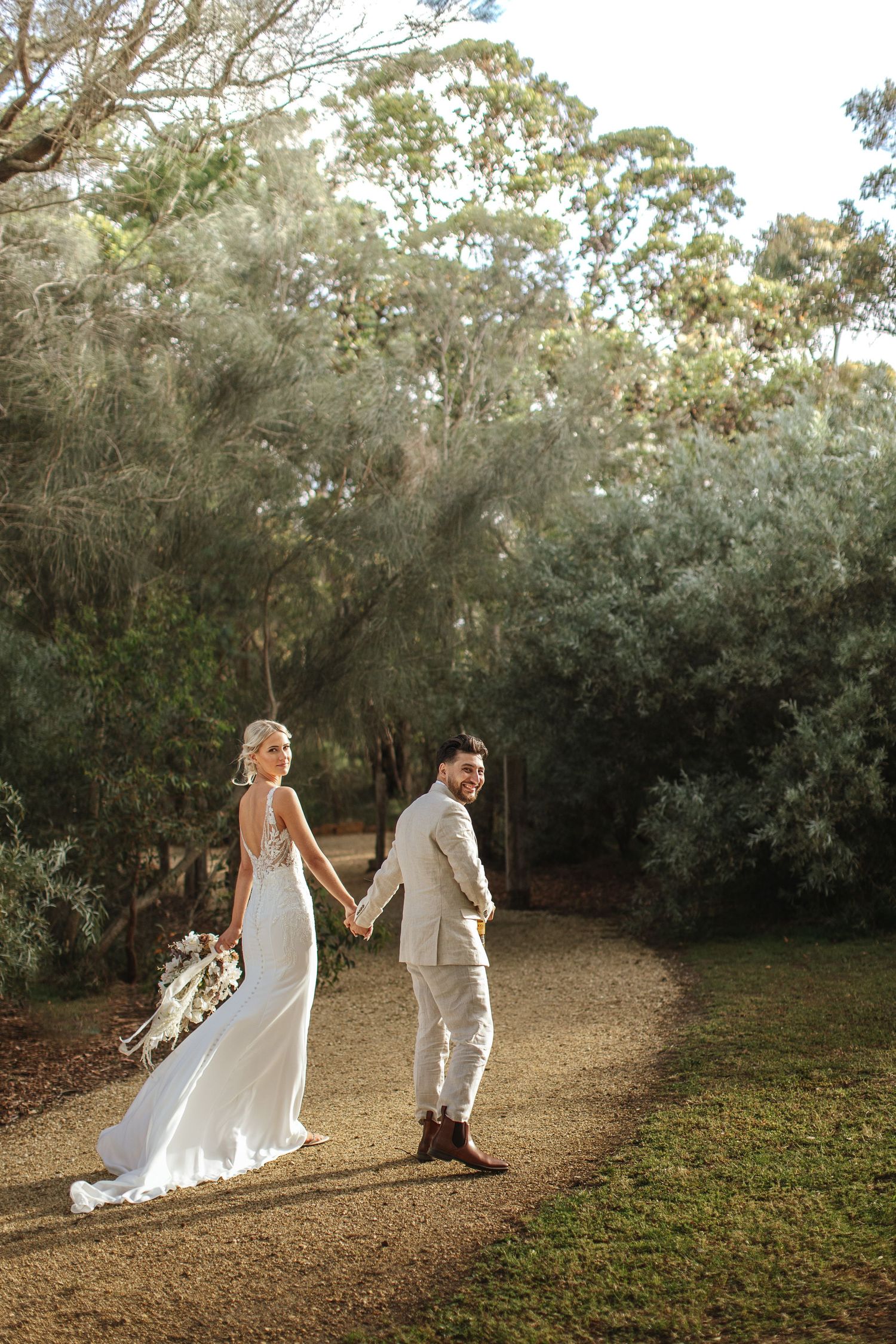 Couple in wedding attire walking hand in hand along forest path at sunset.