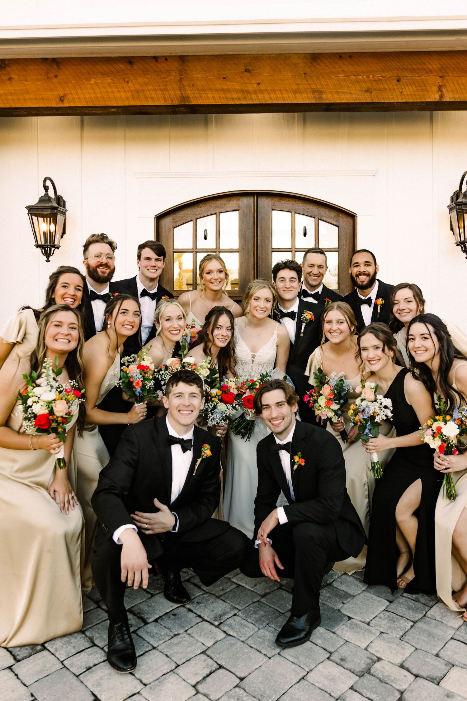 Large wedding party in formal attire poses together outside venue with arched doorway and lanterns.