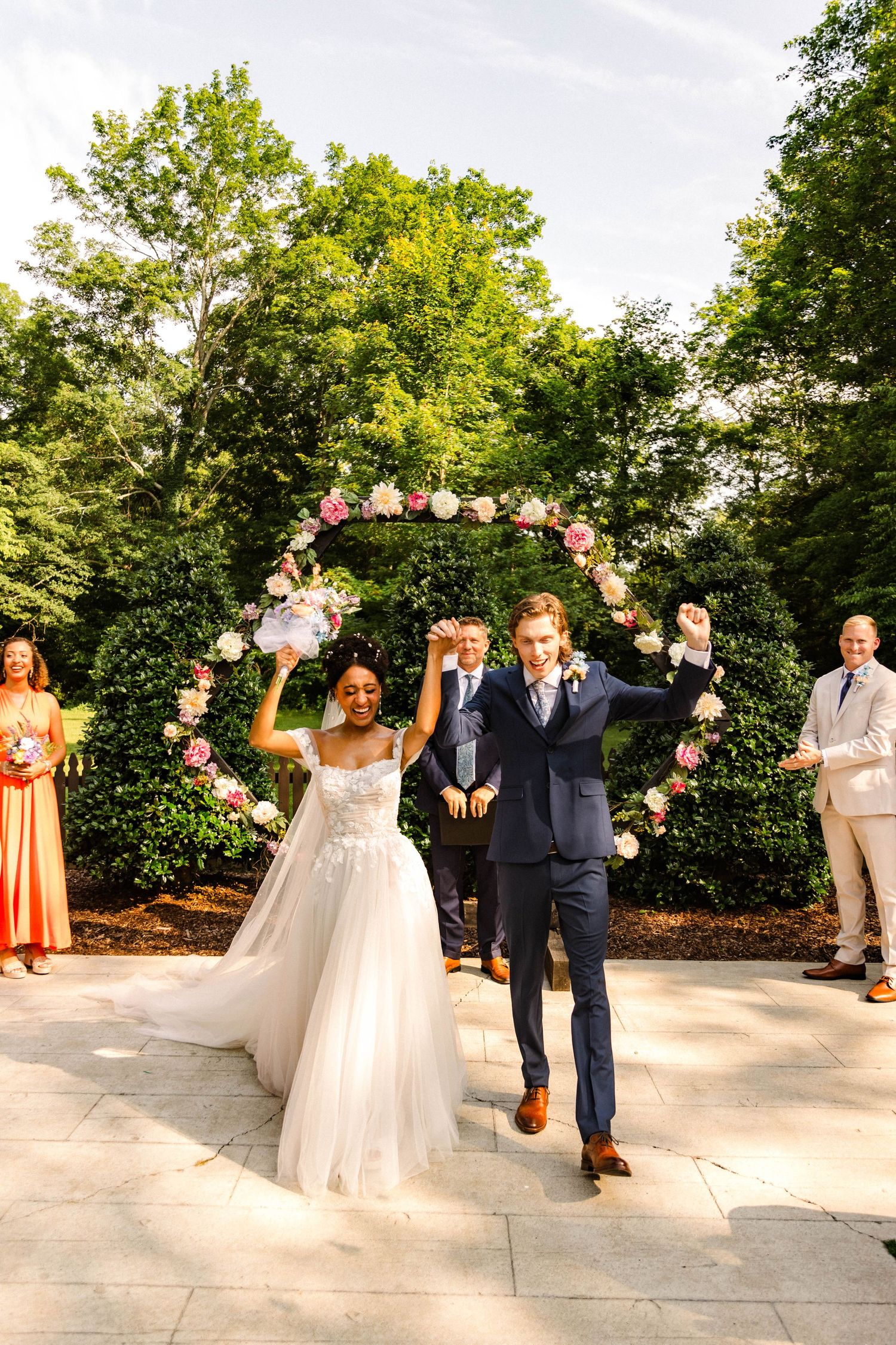 Jubilant couple celebrates under flower-adorned arch during outdoor garden ceremony.