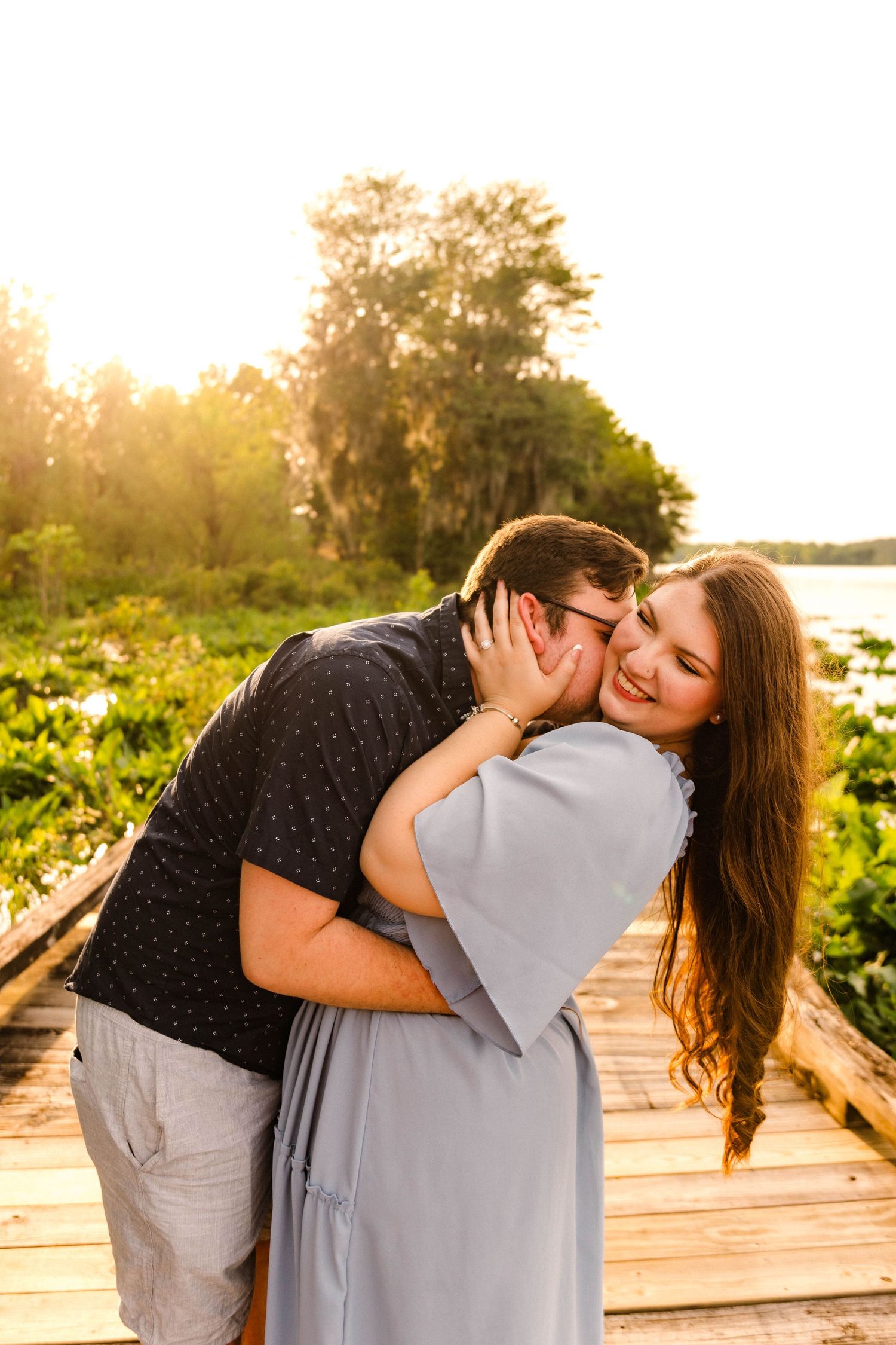 Romantic couple embracing and kissing on a wooden boardwalk during golden hour sunset.