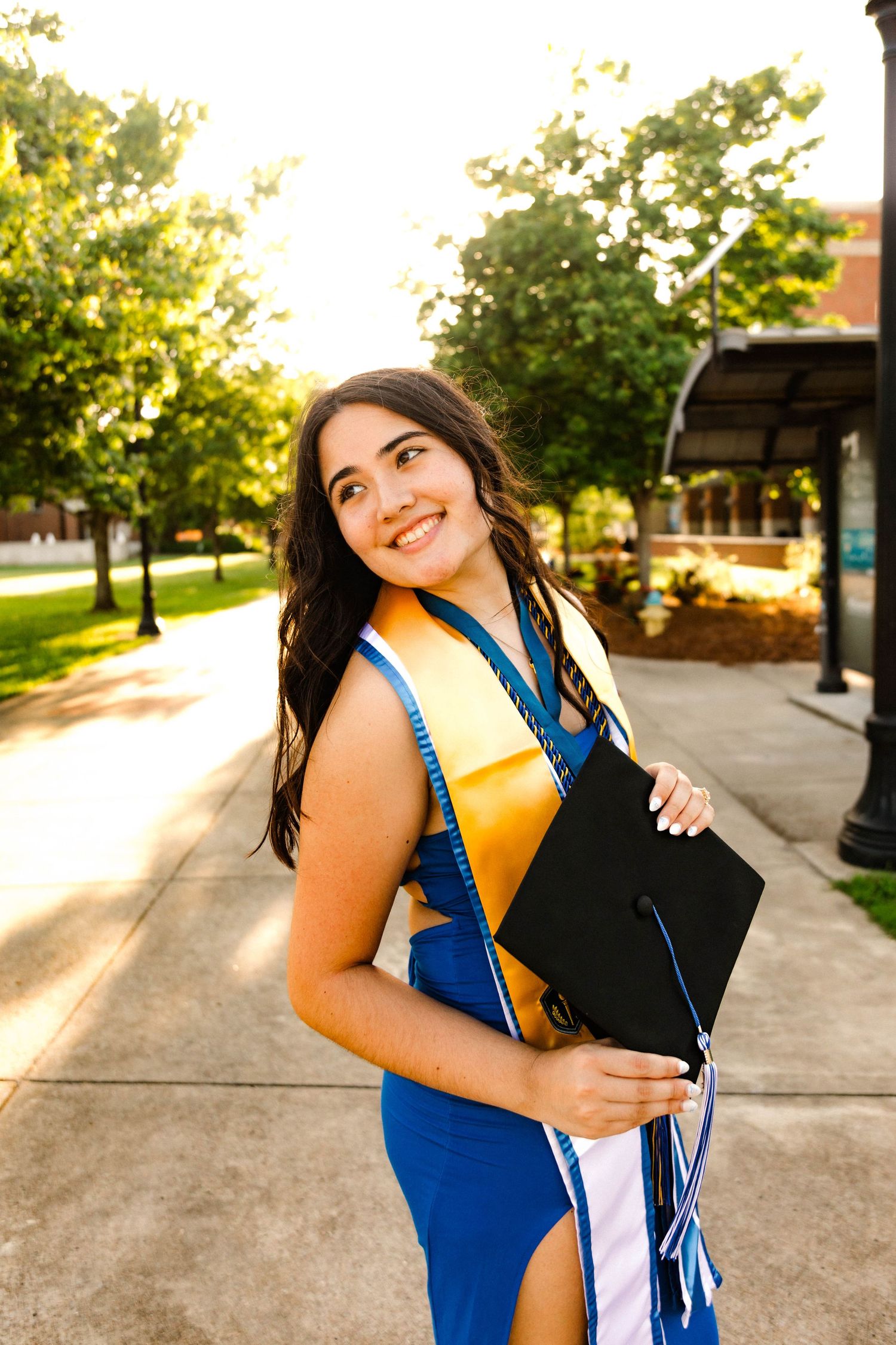 Graduate in blue regalia and gold sash poses with diploma on campus during sunset.