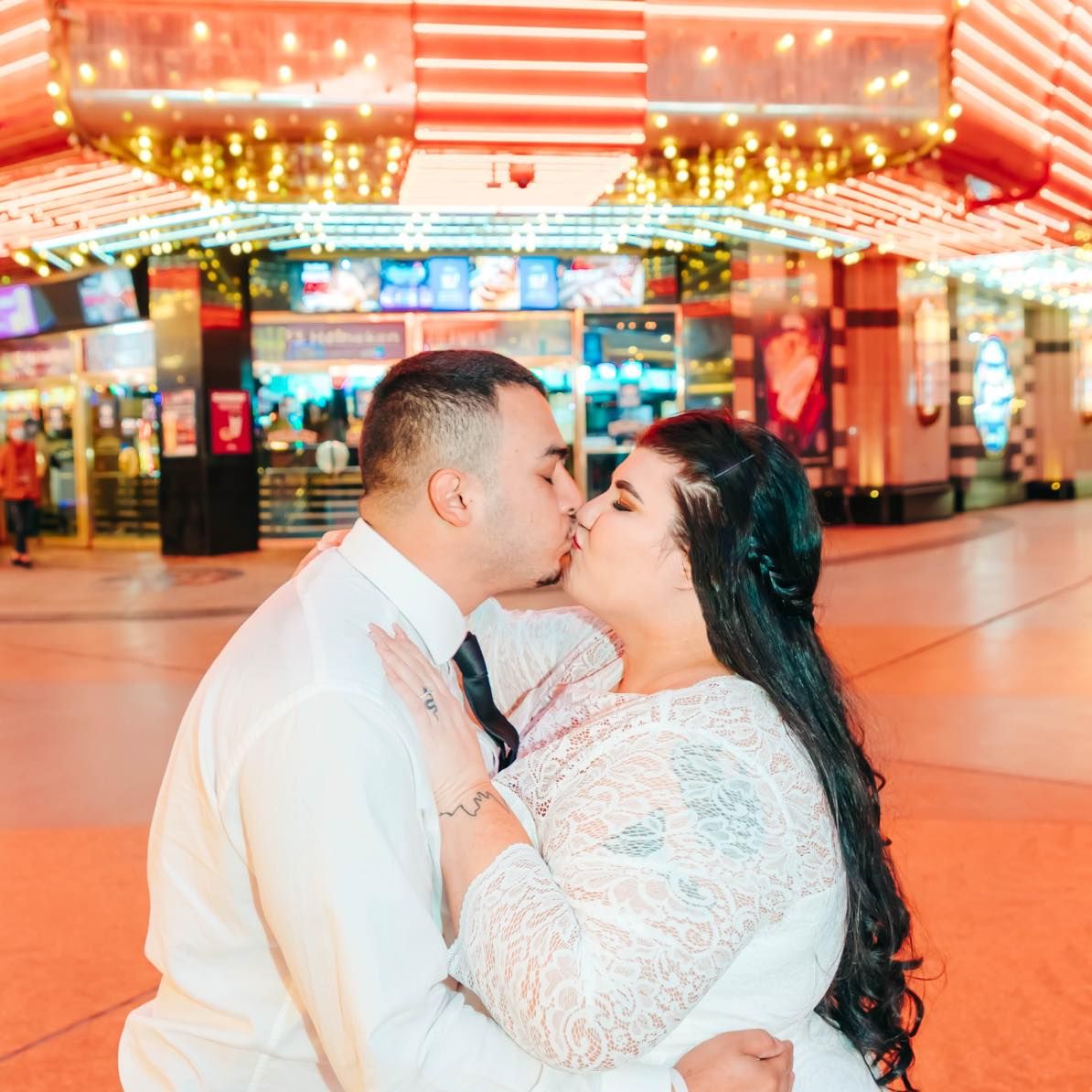 Two people share a tender kiss in front of an illuminated storefront with red and orange lighting.