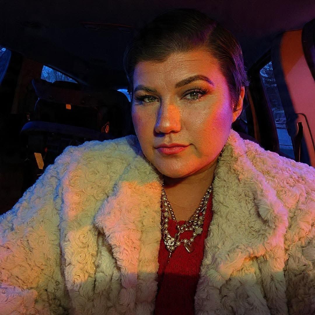 Young woman in white faux fur coat and statement necklace sitting in a car with warm ambient lighting.