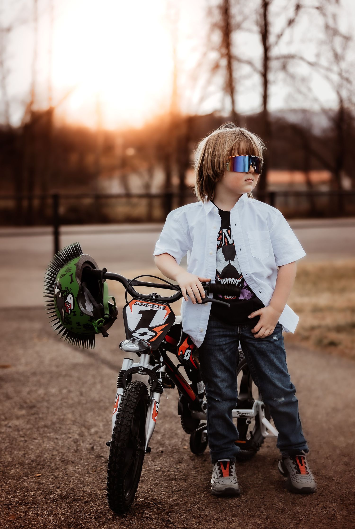 A young person in sunglasses and white shirt stands with a dirt bike at sunset against bare trees in the background.
