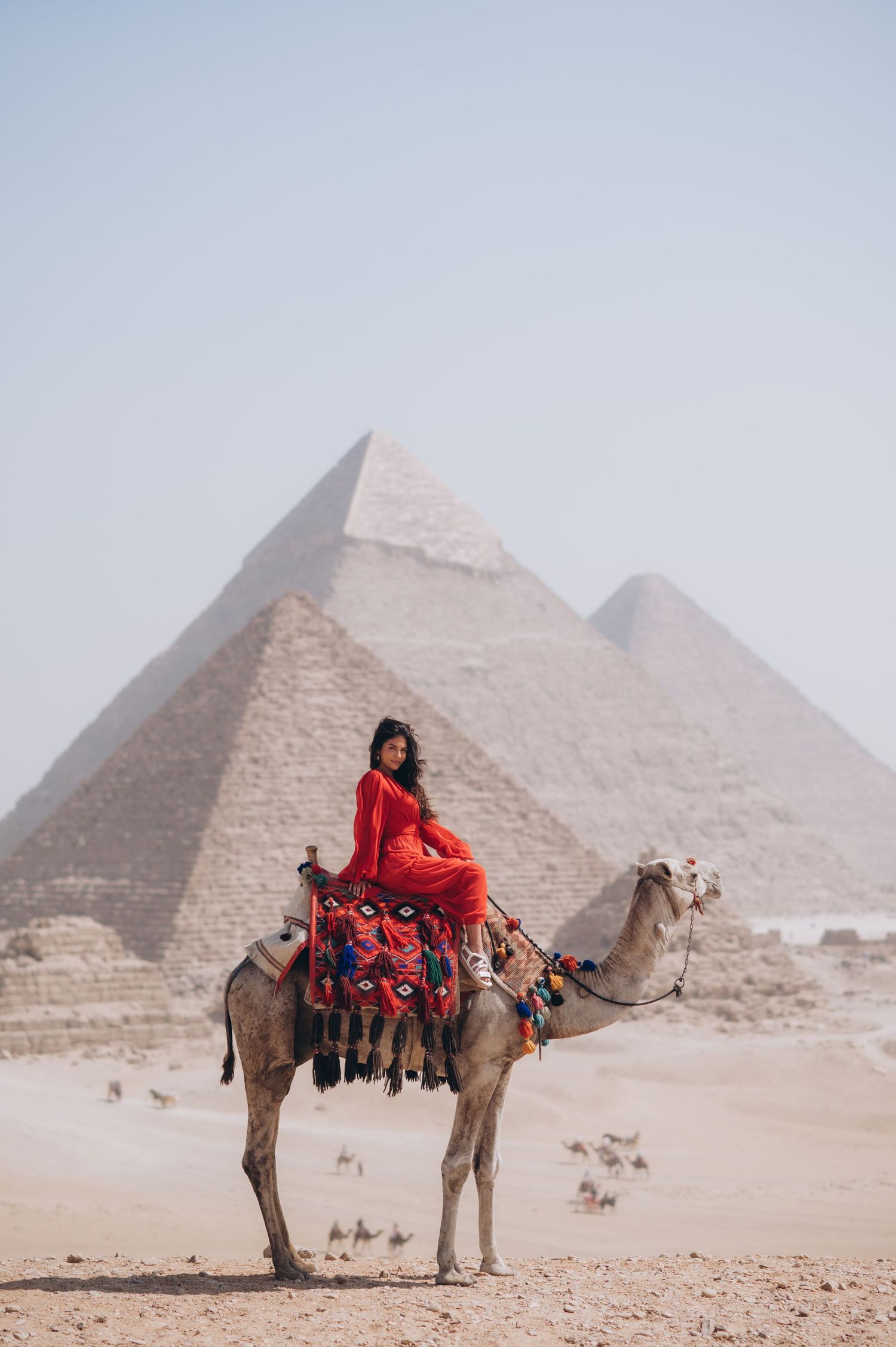 Person in flowing red dress rides decorated camel with pyramids of Giza in background.
