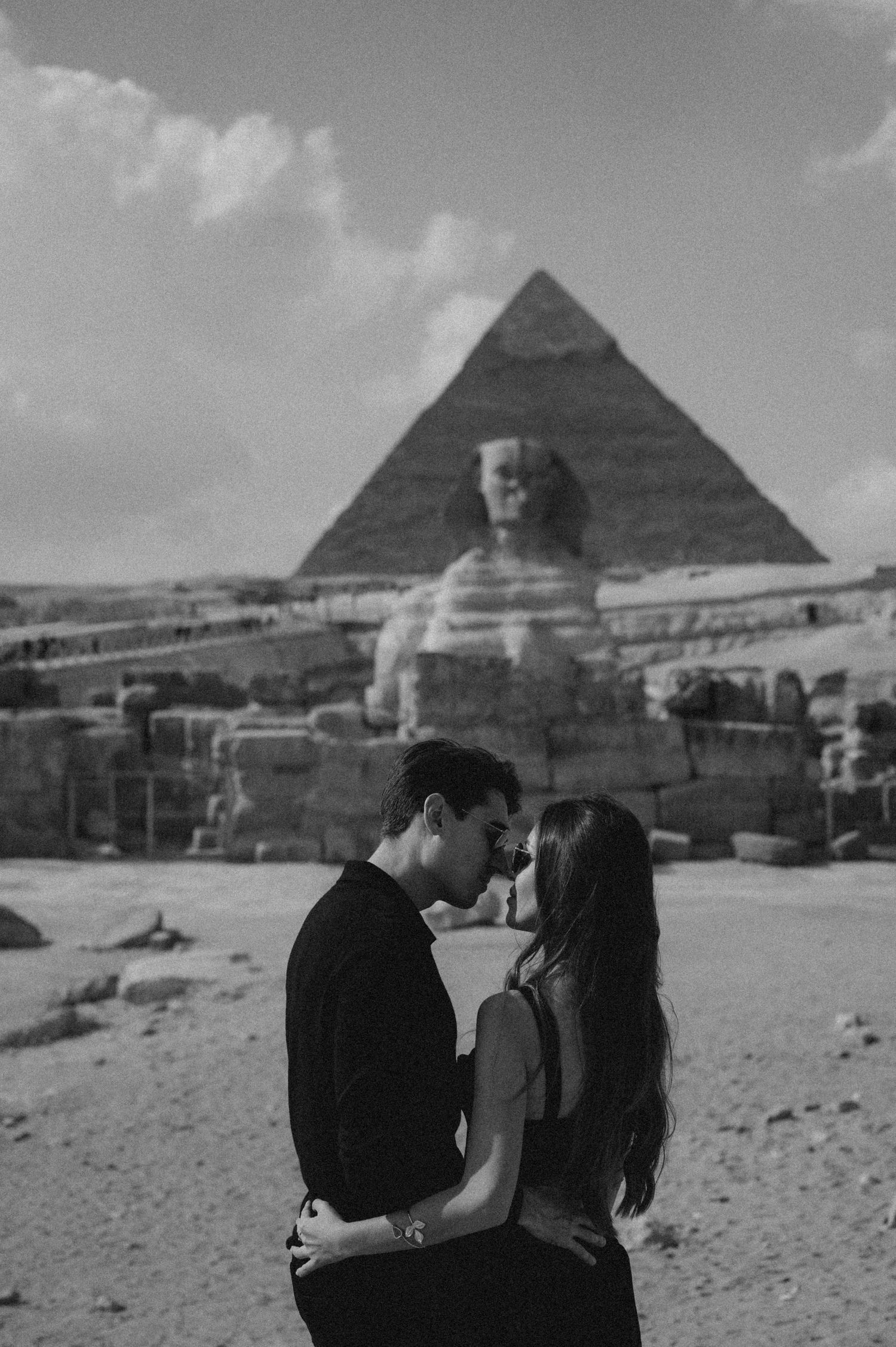 Two figures embrace against a backdrop of the ancient Egyptian pyramids in Giza in black and white photography.