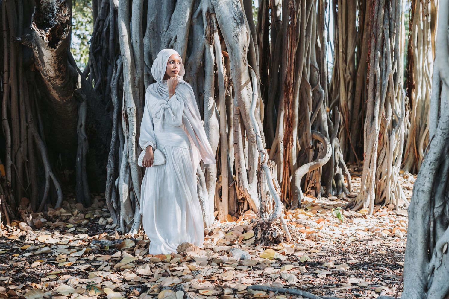 A woman in a flowing white dress poses gracefully among tall banyan trees in a dreamy outdoor setting.