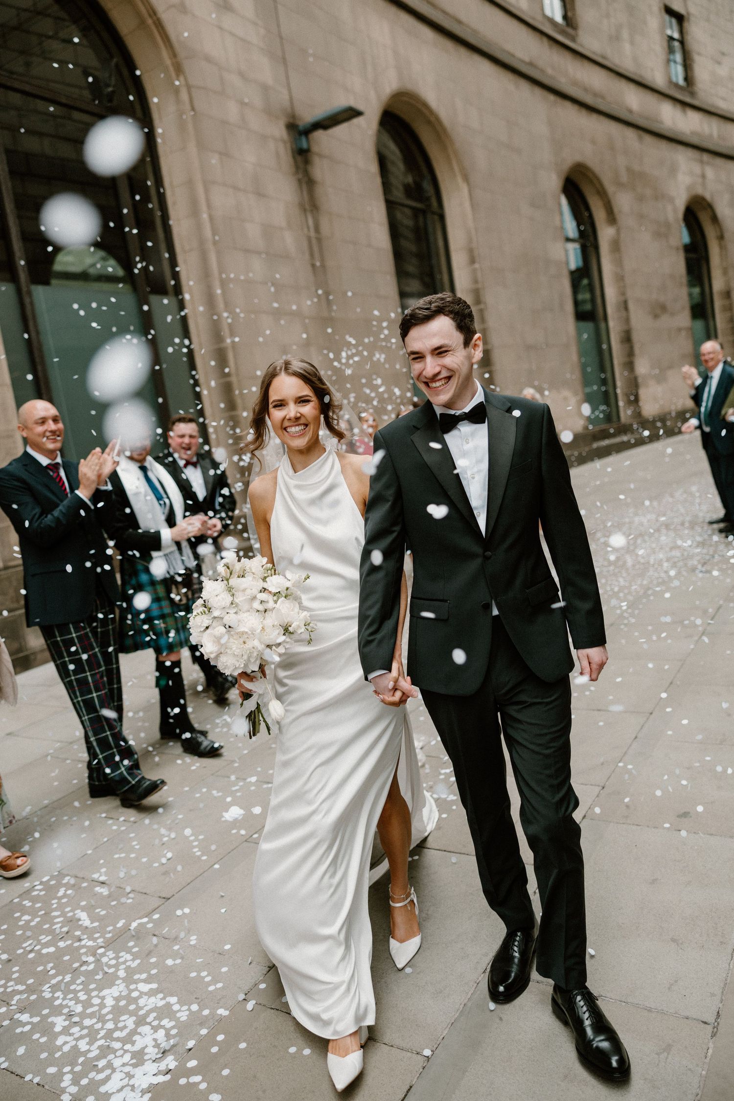 A newlywed couple exits their wedding venue while guests shower them with white confetti on a city street.