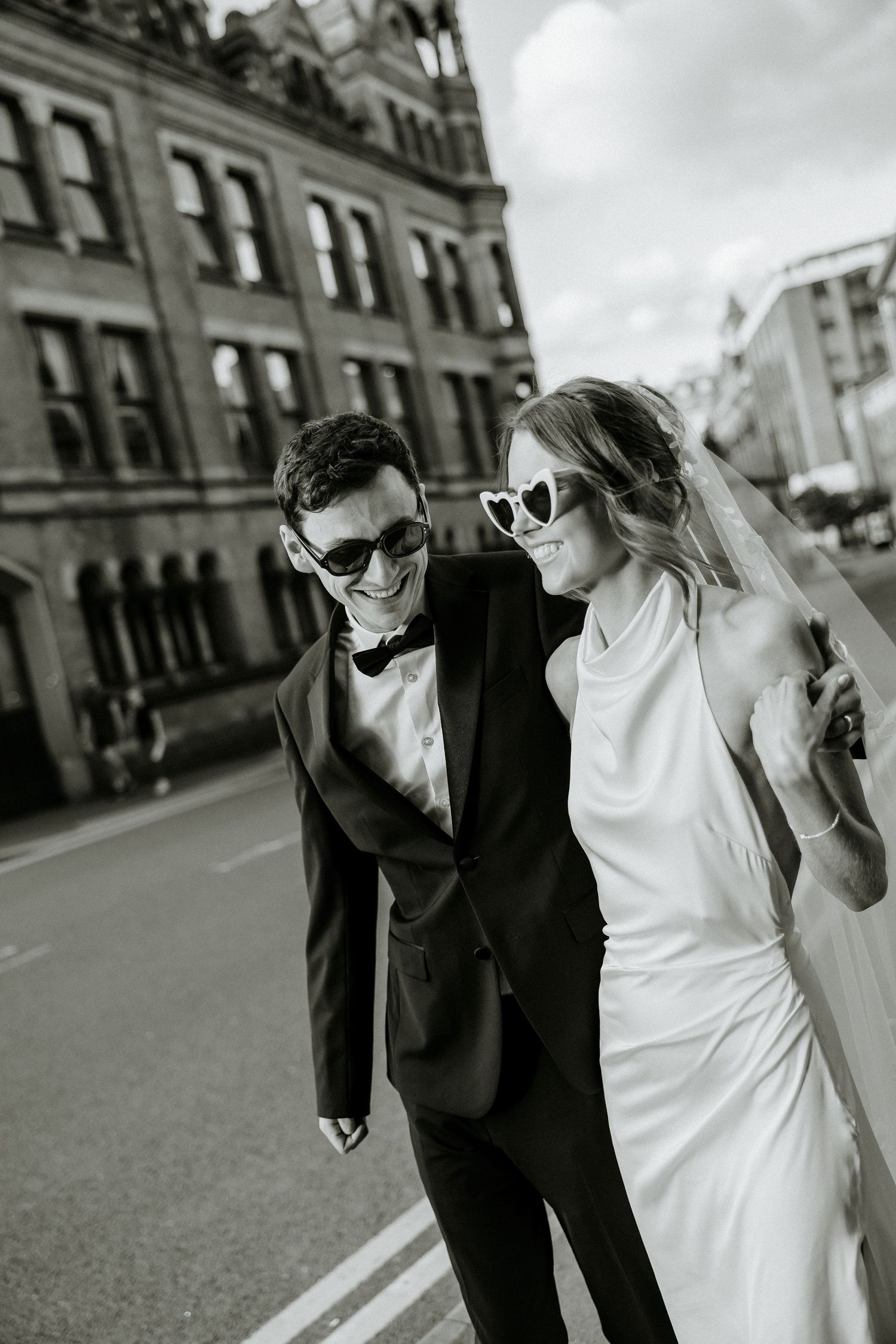 A couple wearing wedding attire walks together along a cobblestone street in a candid black and white photo series.