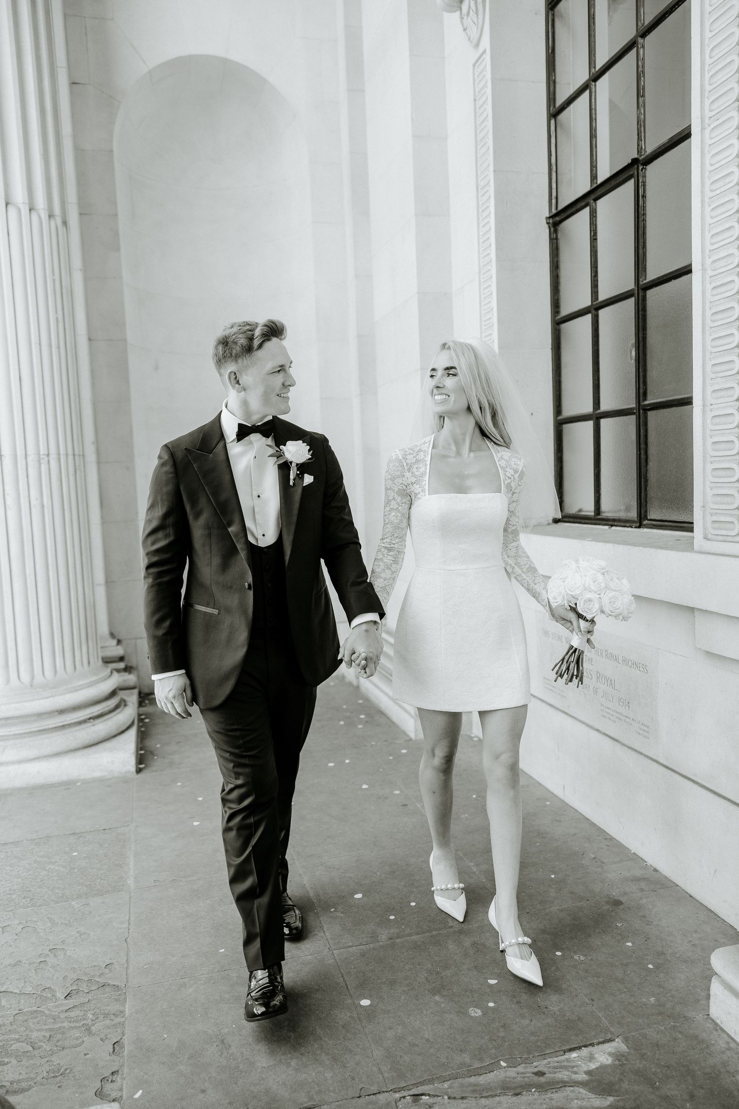 A bride in a short white dress and groom in black tuxedo walk together next to classic white columns in this retro photo.