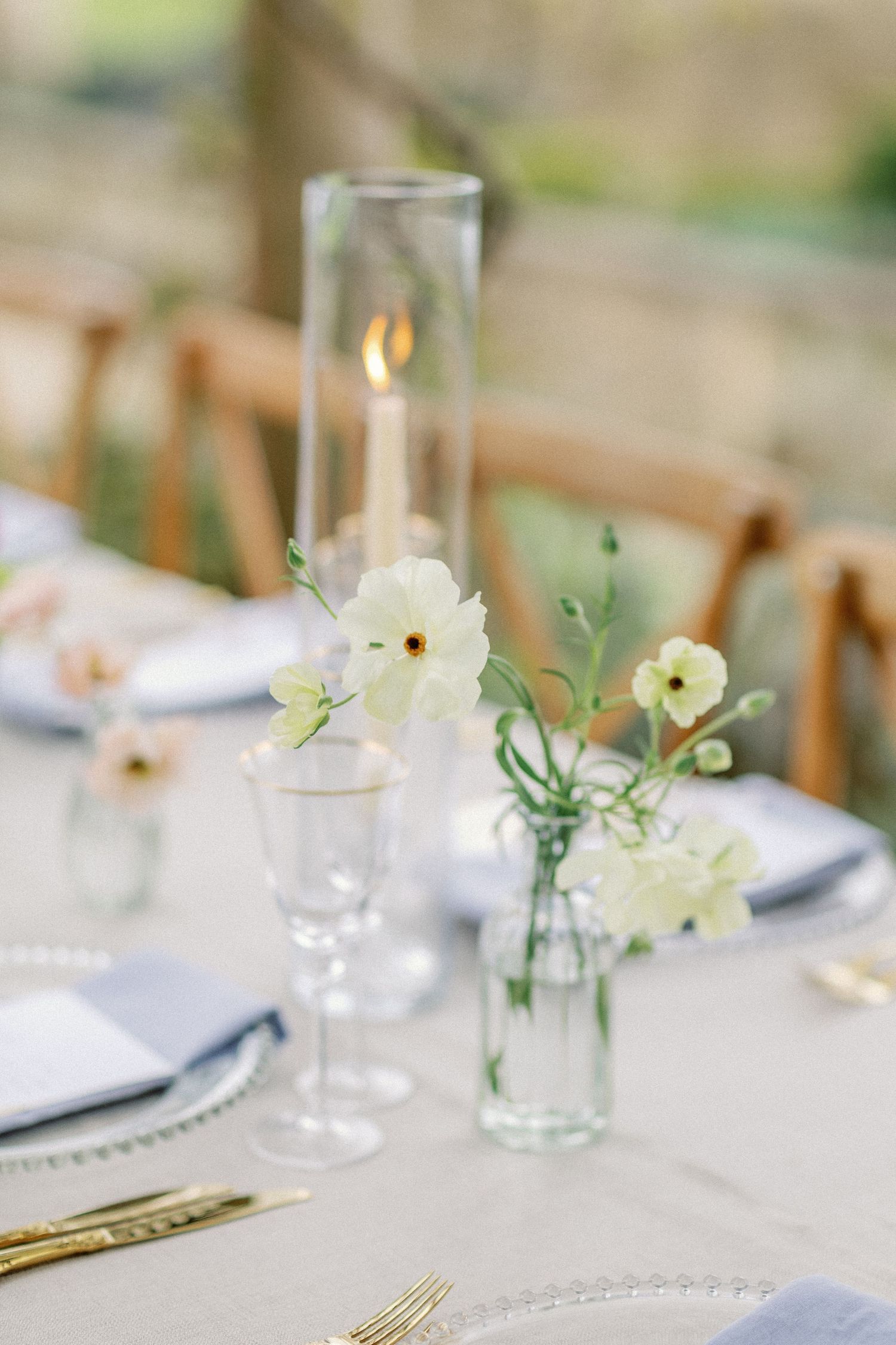 Delicate table setting with glass candleholder and white flowers in small vases.