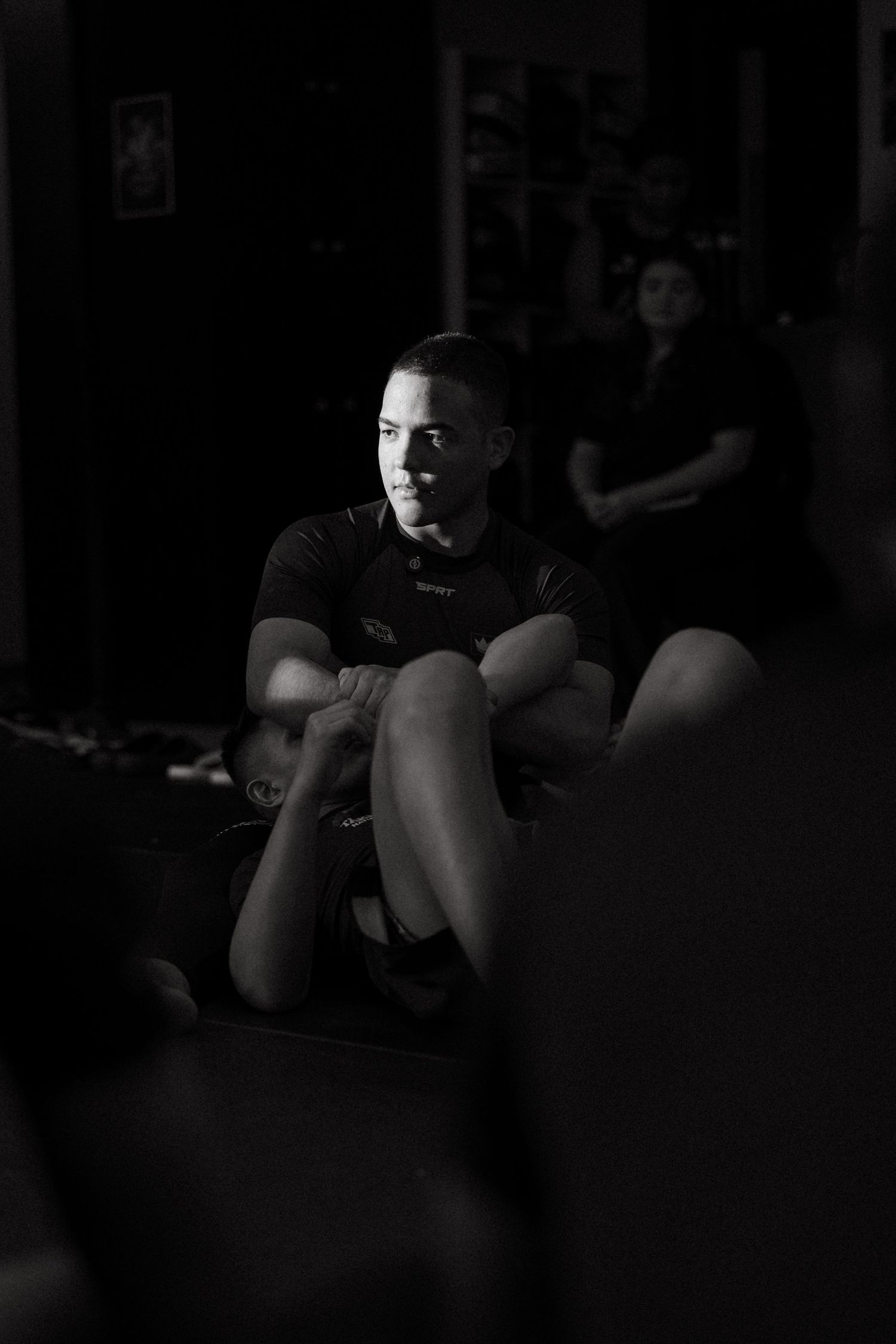 Black and white photo of a focused young male athlete sitting cross-legged in a dark gym environment.