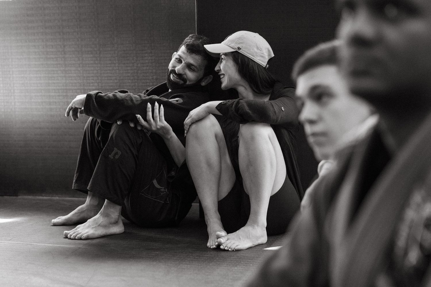 Two martial artists in gi uniforms rest on a gym floor during a Brazilian jiu-jitsu training session.