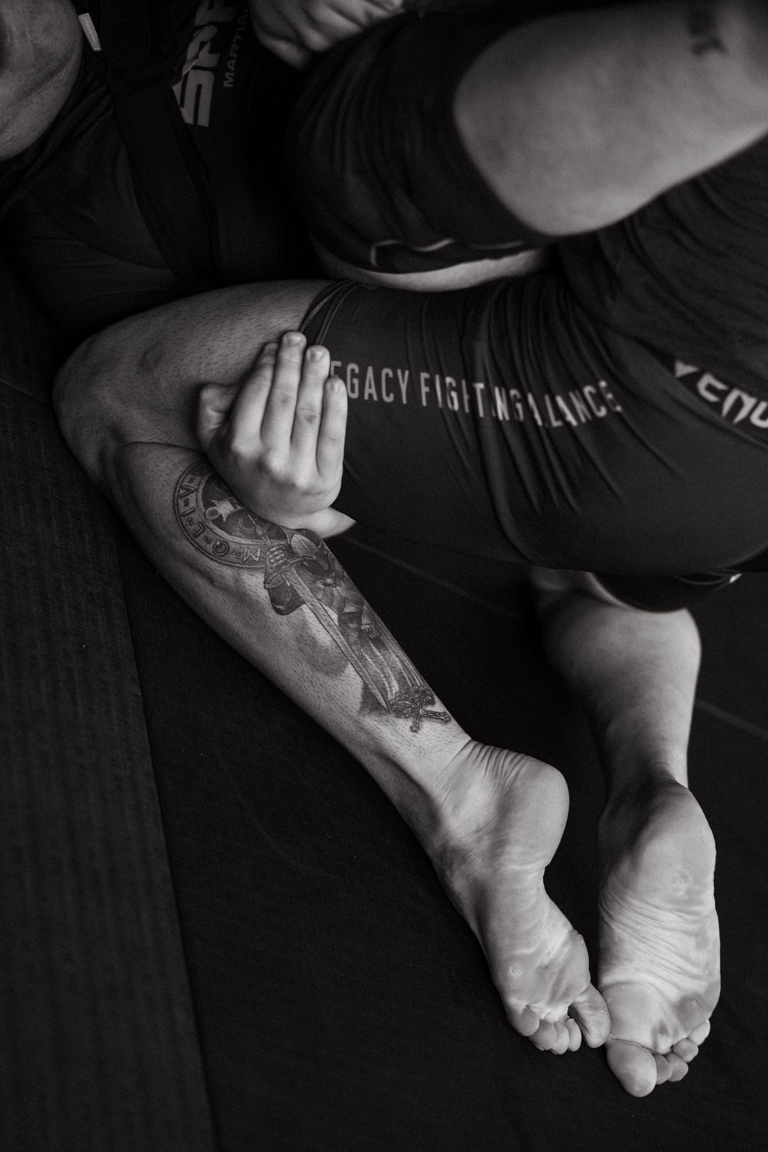 Black and white photo of MMA fighters grappling, showing tattooed arm and bare feet on a mat.