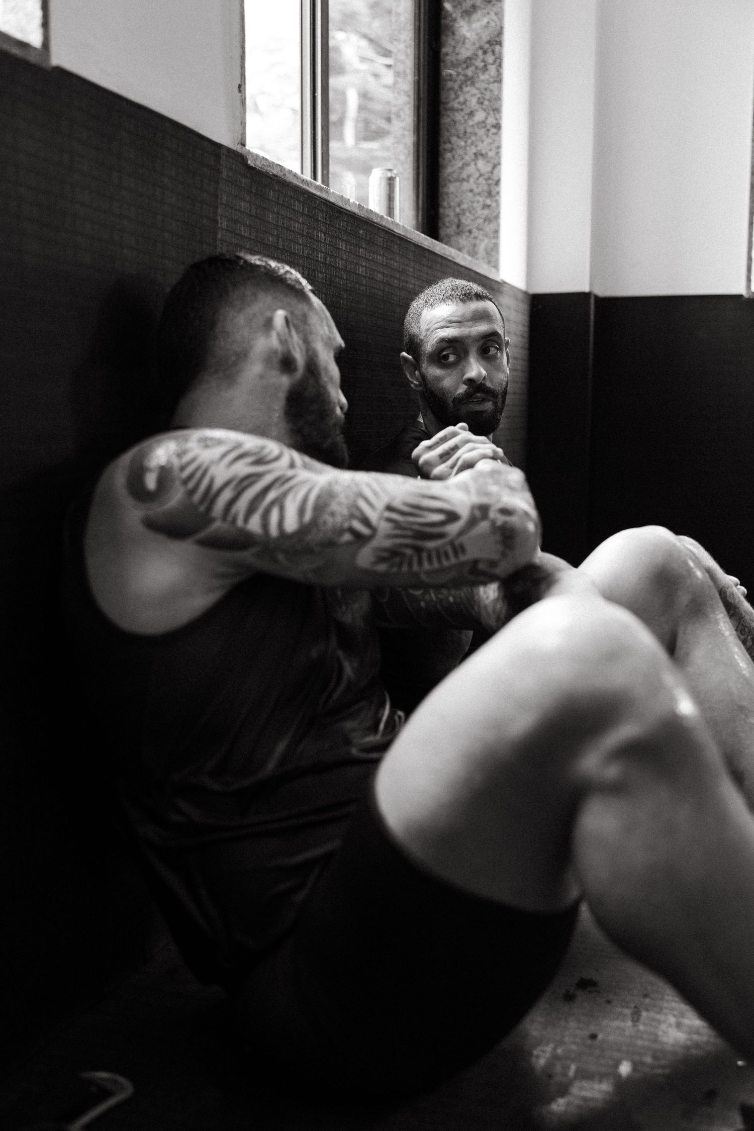 Two tattooed muscular men resting against a gym wall in a black and white photo.