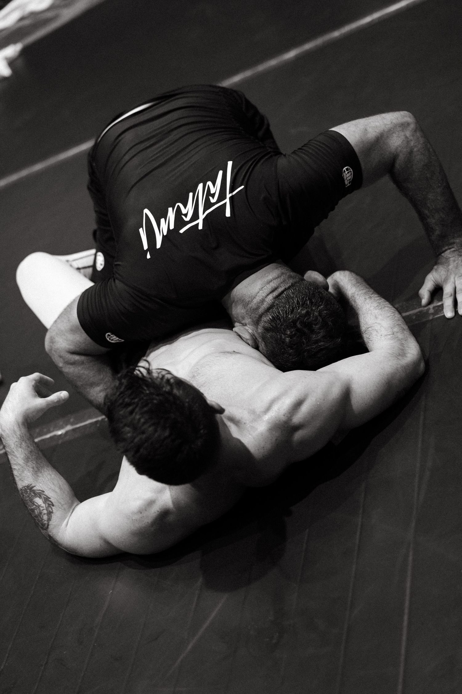Two BJJ athletes grappling on a mat, one wearing a black Tatami rashguard, in black and white.