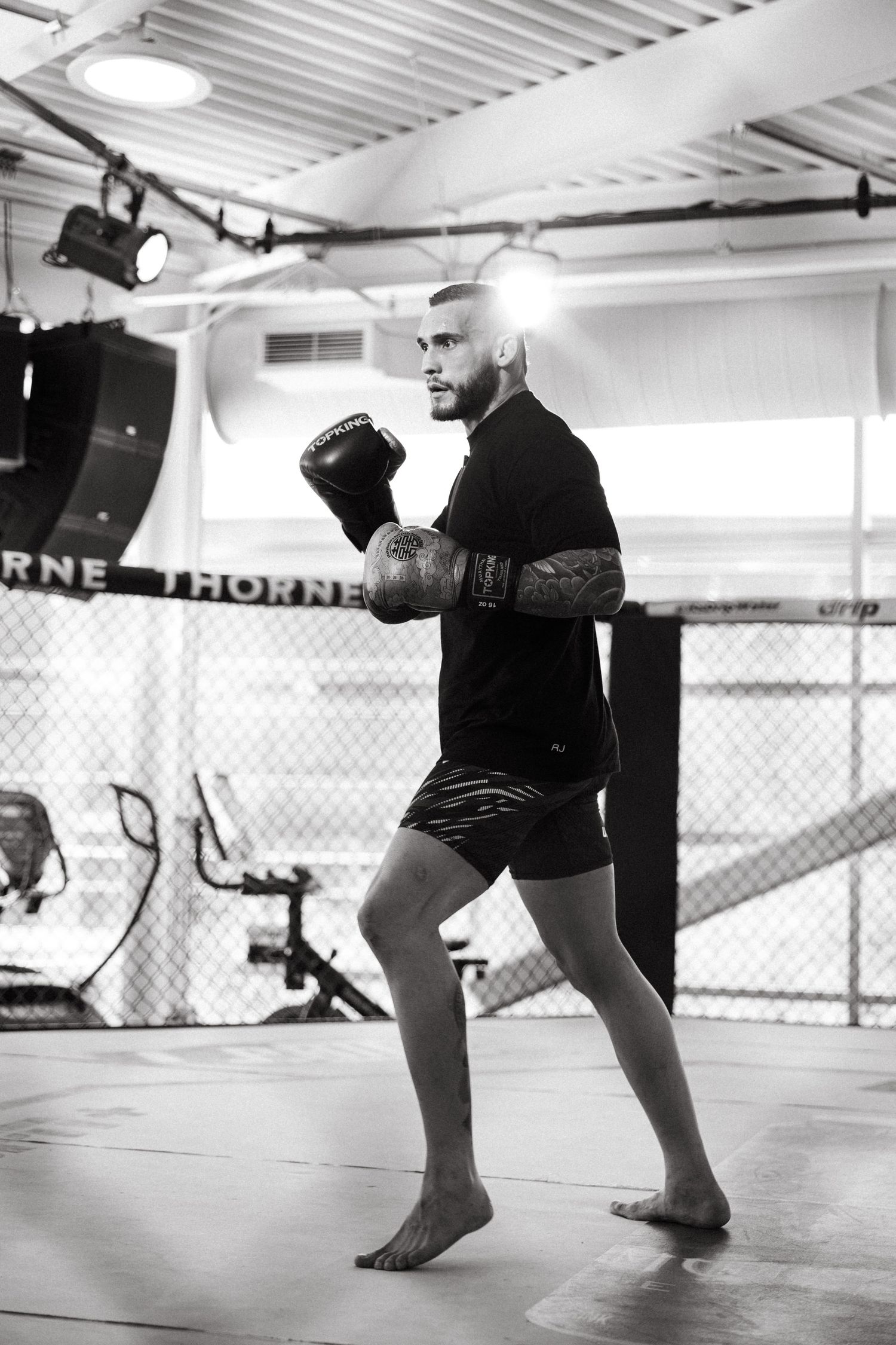 Black and white photo of a male MMA fighter in boxing gloves training in a gym with an octagon cage in the background.