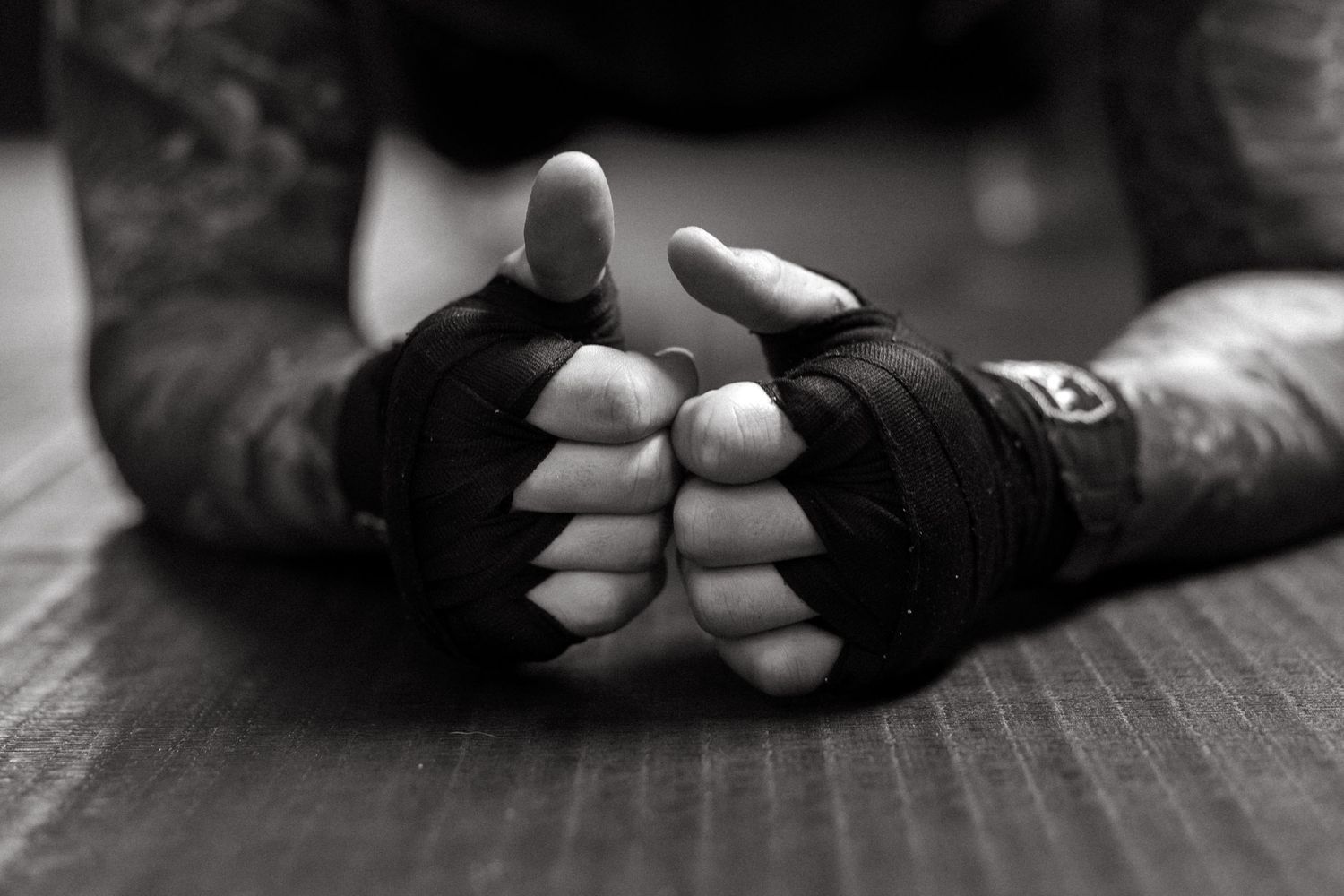 Black and white close-up of tattooed hands wrapped in black boxing wraps resting on a gym mat.