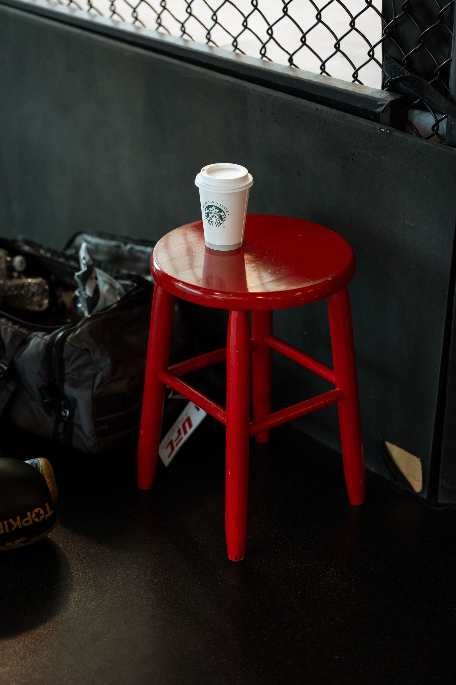 A white Starbucks cup sits on a small red wooden stool against a dark wall with chain-link fencing.