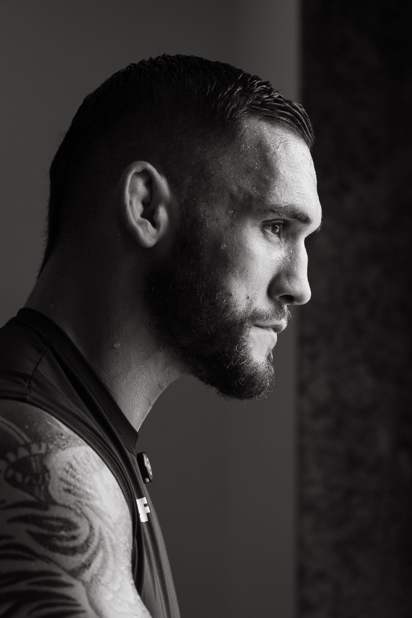 Black and white profile portrait of a tattooed man with beard and slicked hair in dramatic lighting.