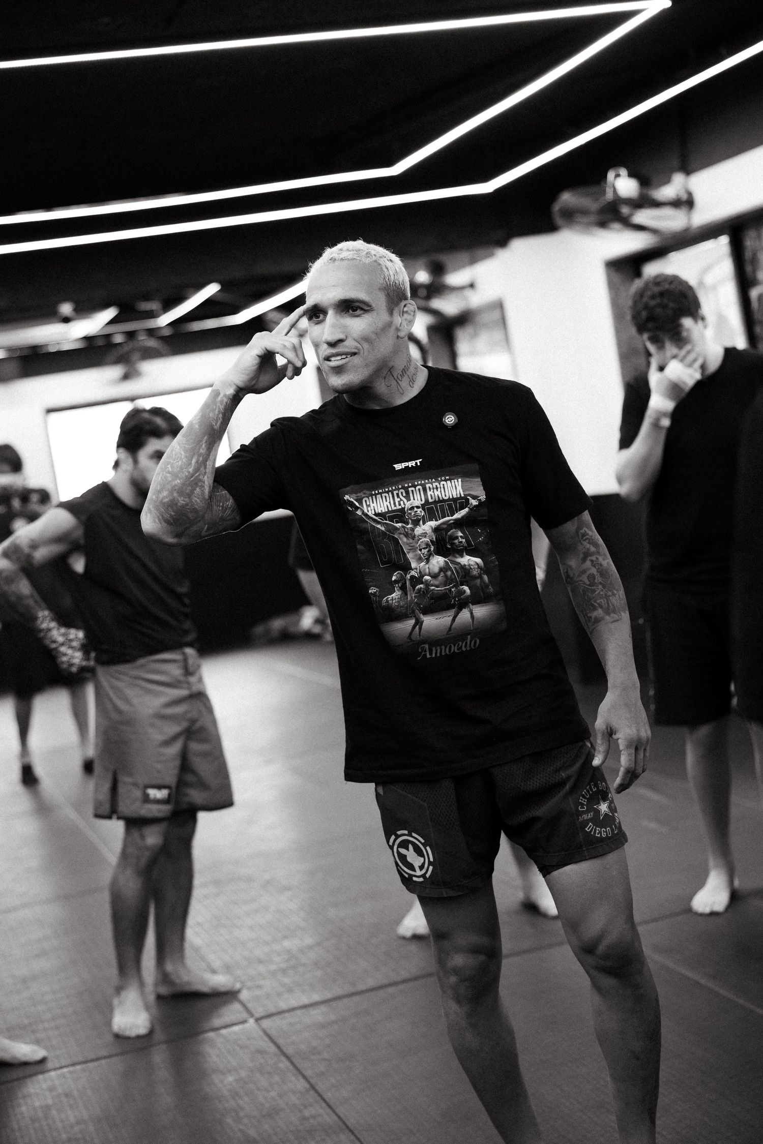 Black and white photo of a tattooed man in MMA shorts and graphic tee at a martial arts gym.