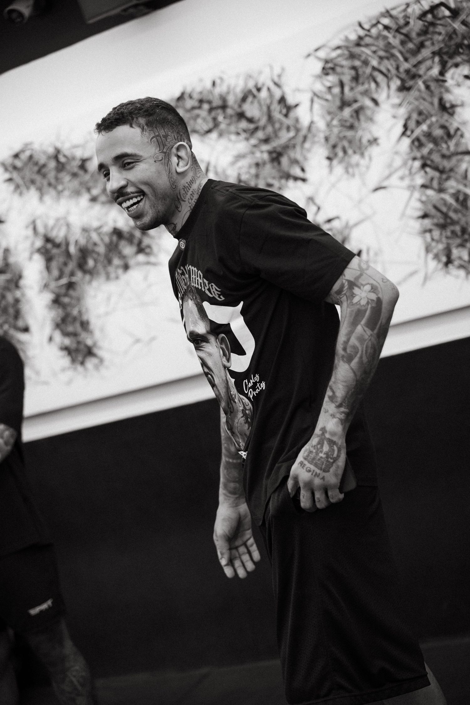 Black and white photo of a tattooed smiling man in a black graphic tee, laughing outdoors near foliage.
