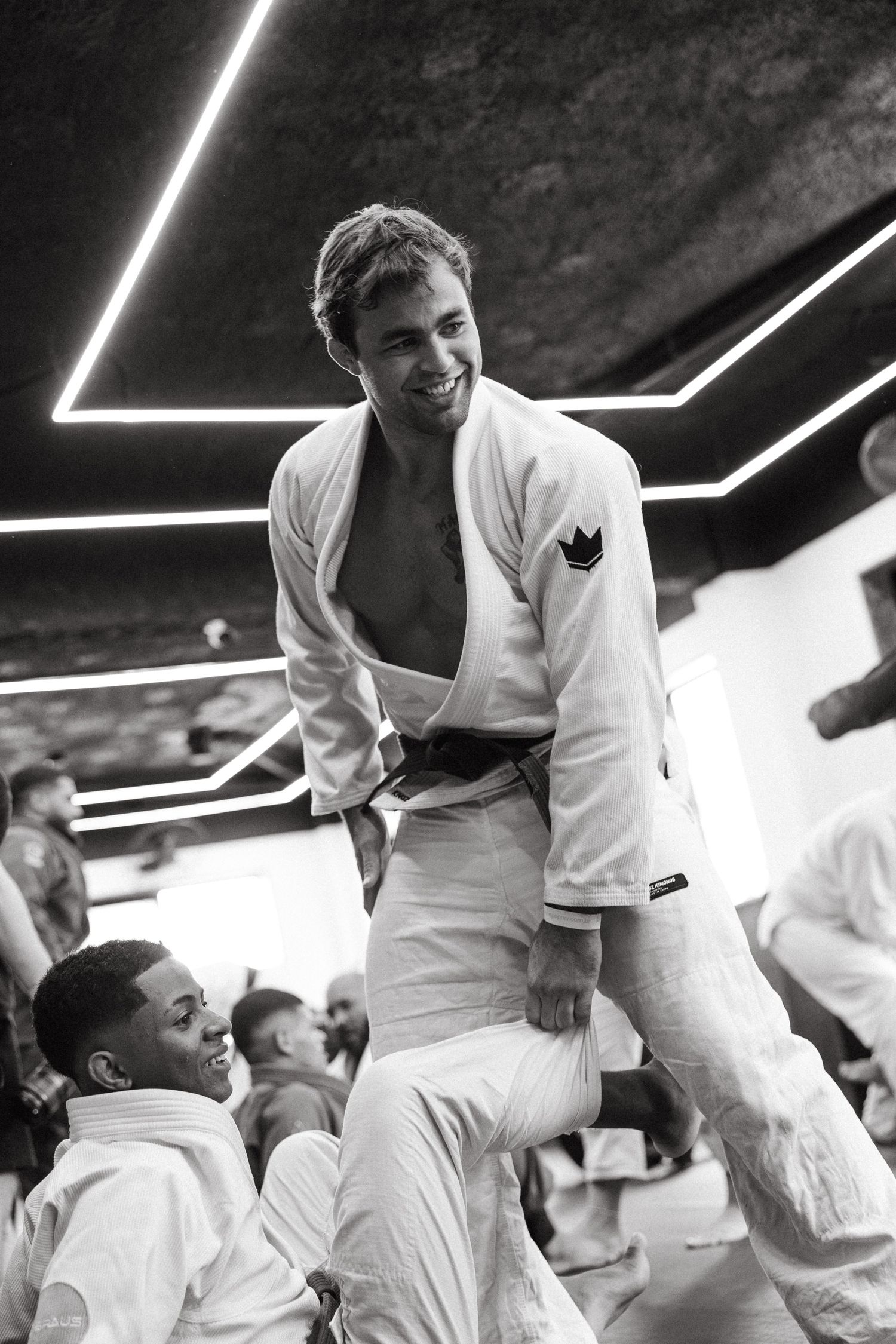 Two men in white BJJ gis celebrate a belt promotion in a martial arts gym with geometric neon lighting.