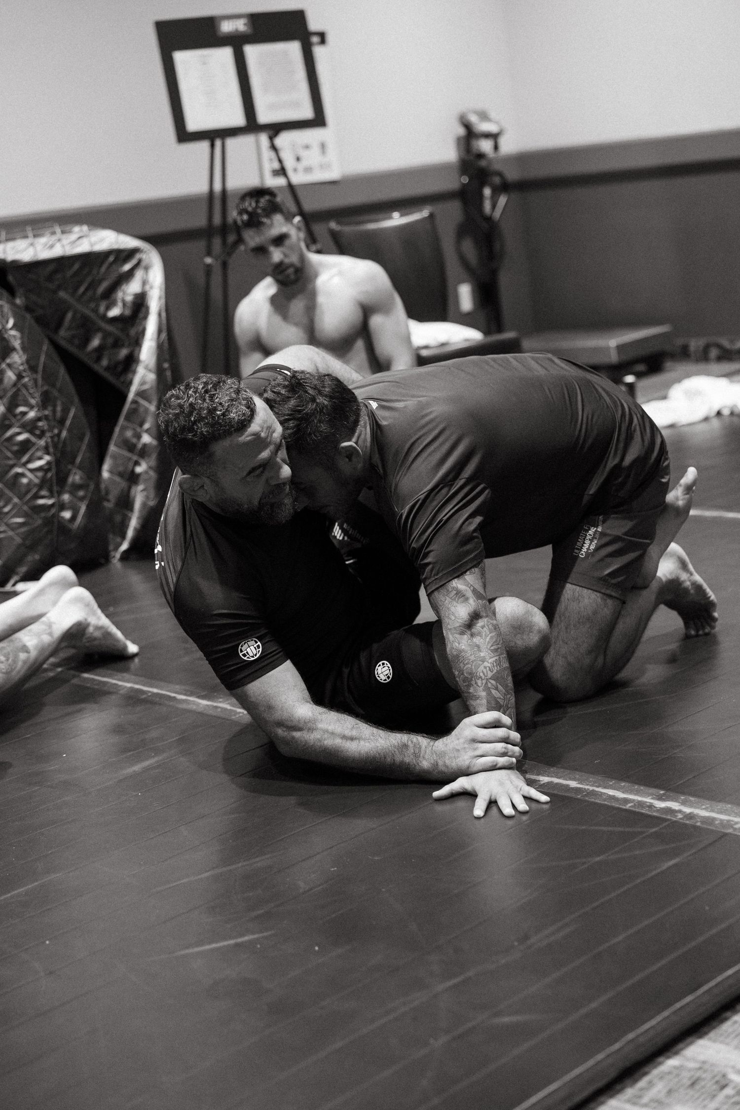 Two athletes practice grappling techniques on a mat in a gym during a Brazilian jiu-jitsu training session.