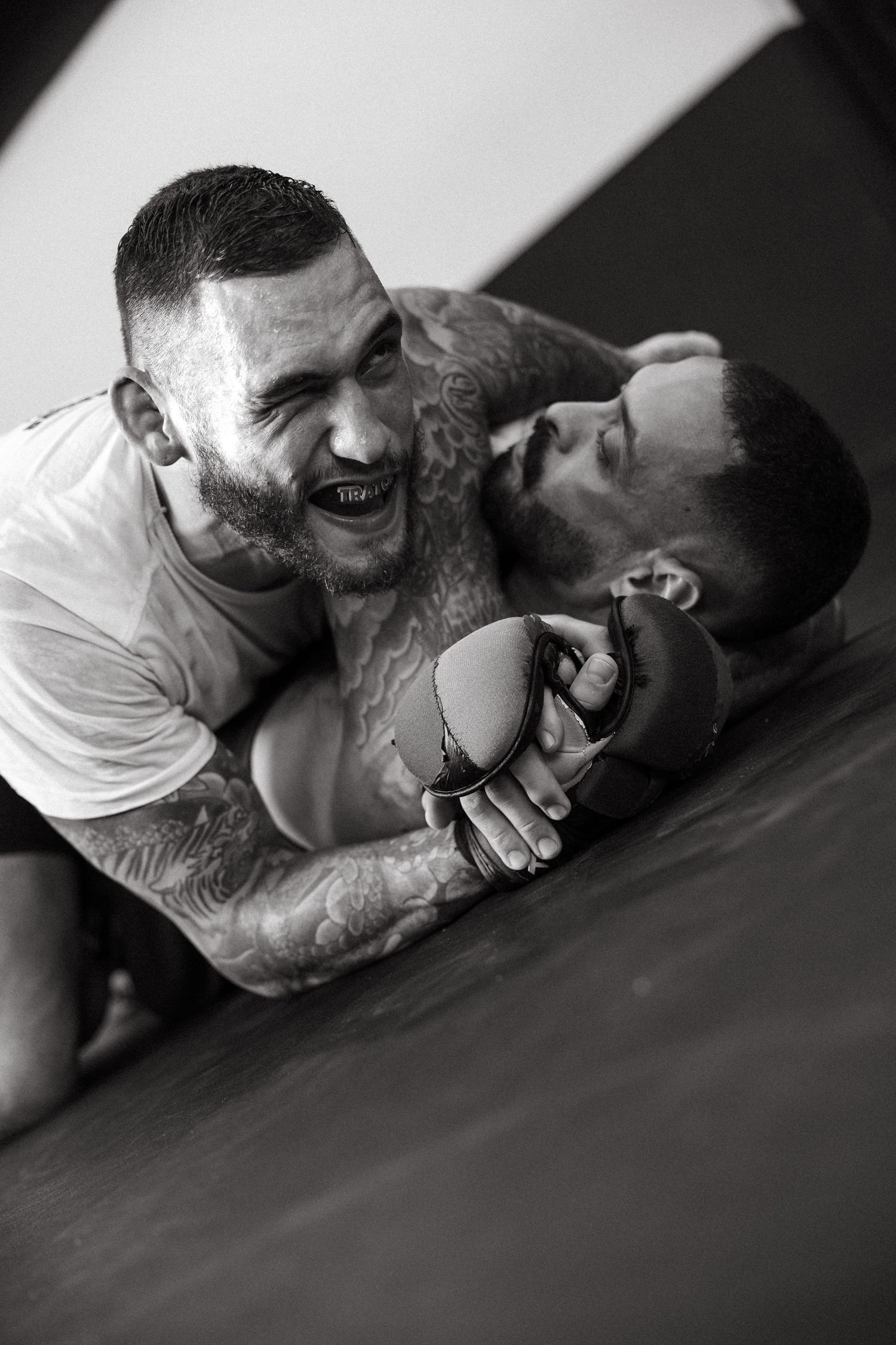 Two tattooed MMA fighters grappling on the mat in an intense ground fight, wearing MMA gloves, black and white photo.