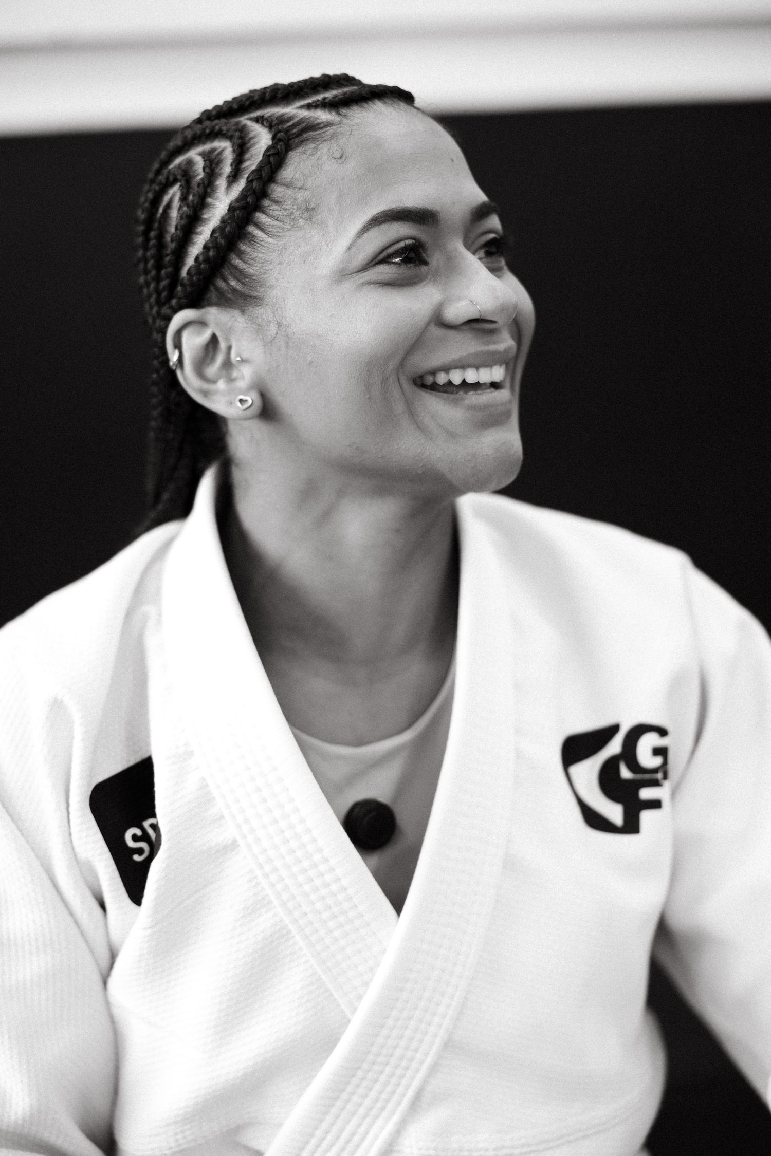 Black and white portrait of a smiling woman in a white judo gi with braided hair and stud earrings.