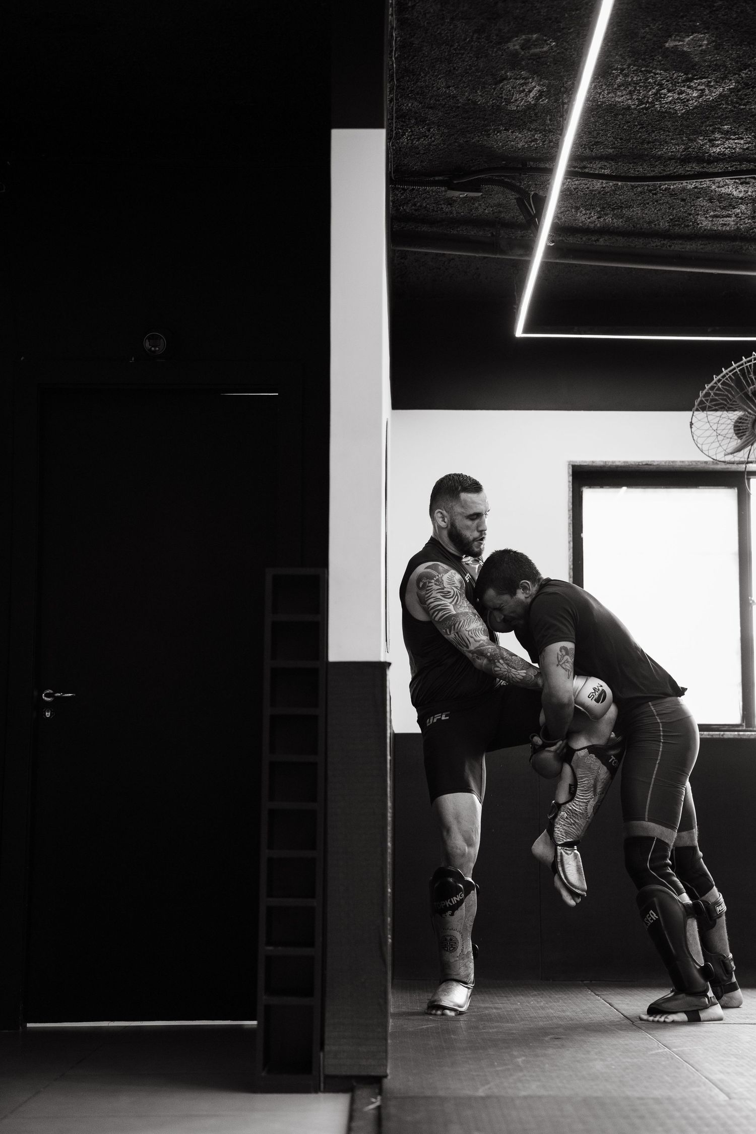 Two men practicing MMA grappling techniques in a dark gym, black and white photography.