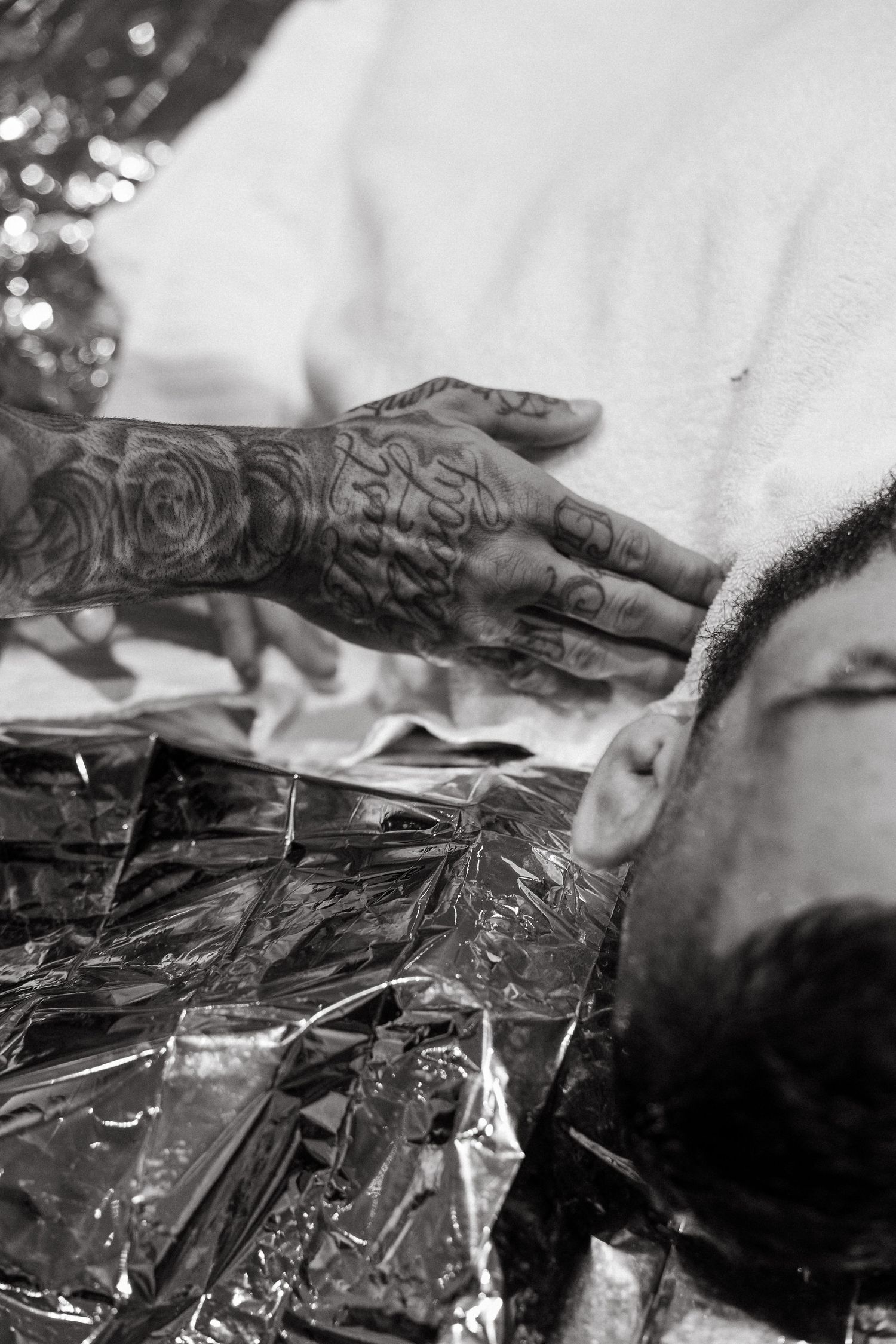 Black and white photo of tattooed hands styling a bearded mans hair in a barbershop.