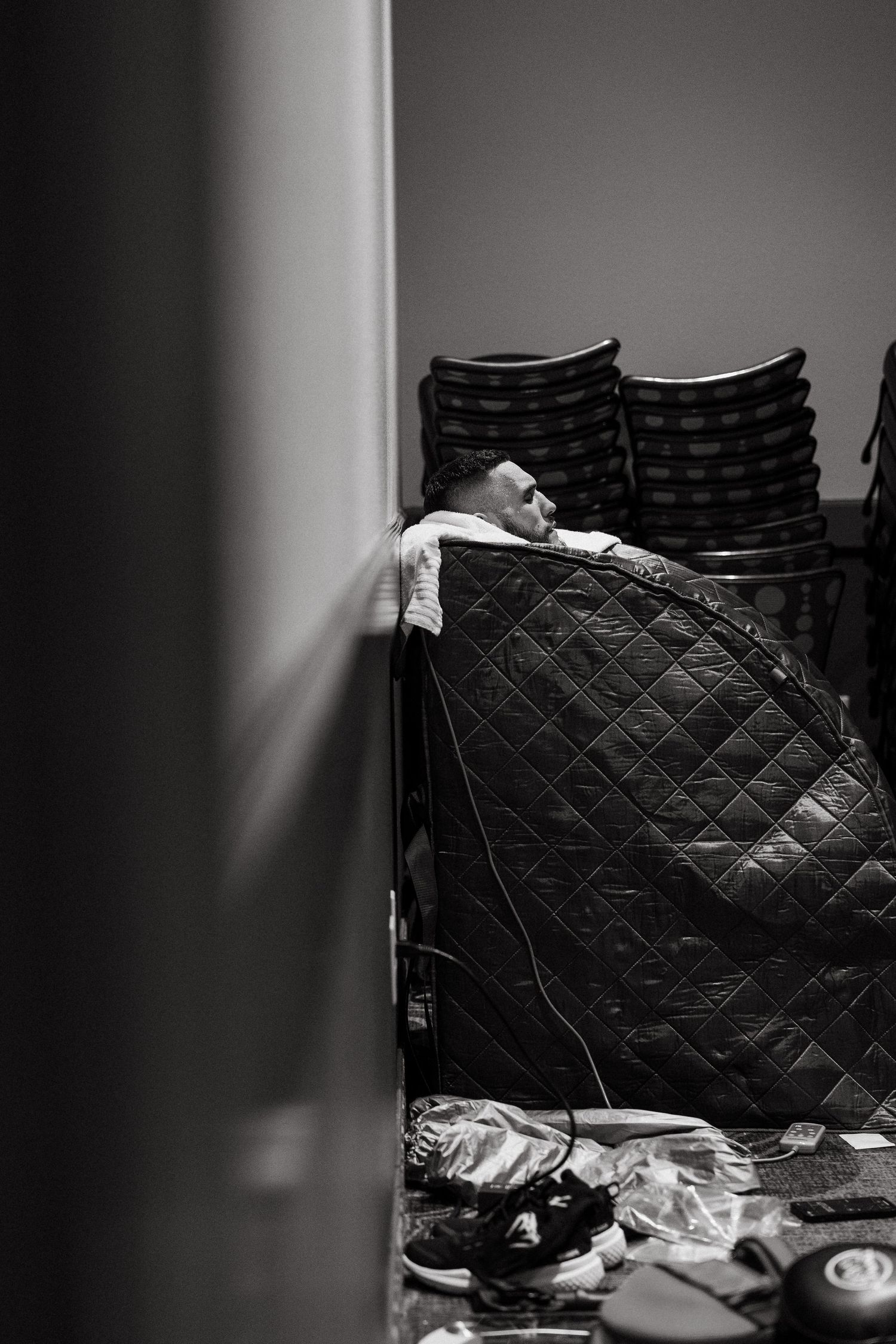 Black and white photo of a person resting behind a quilted chair with stacked chairs and scattered items on the floor.