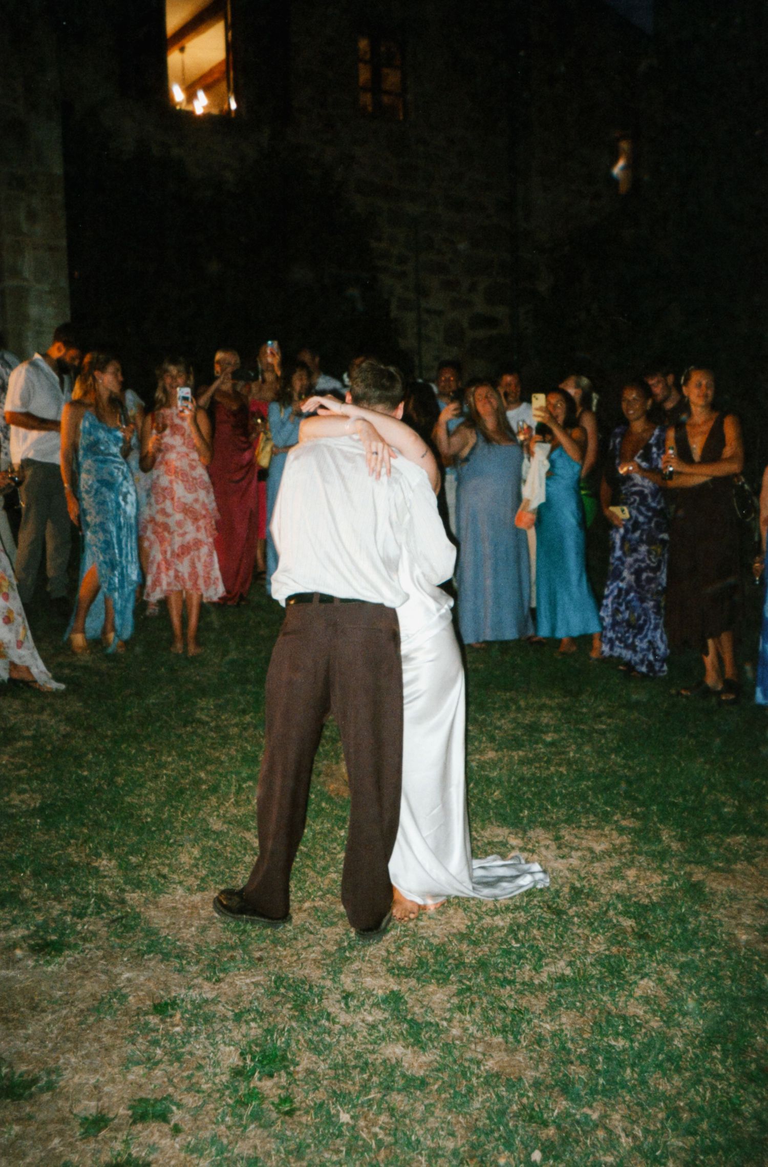 Wedding guests dance together on a grassy lawn at an outdoor evening reception.