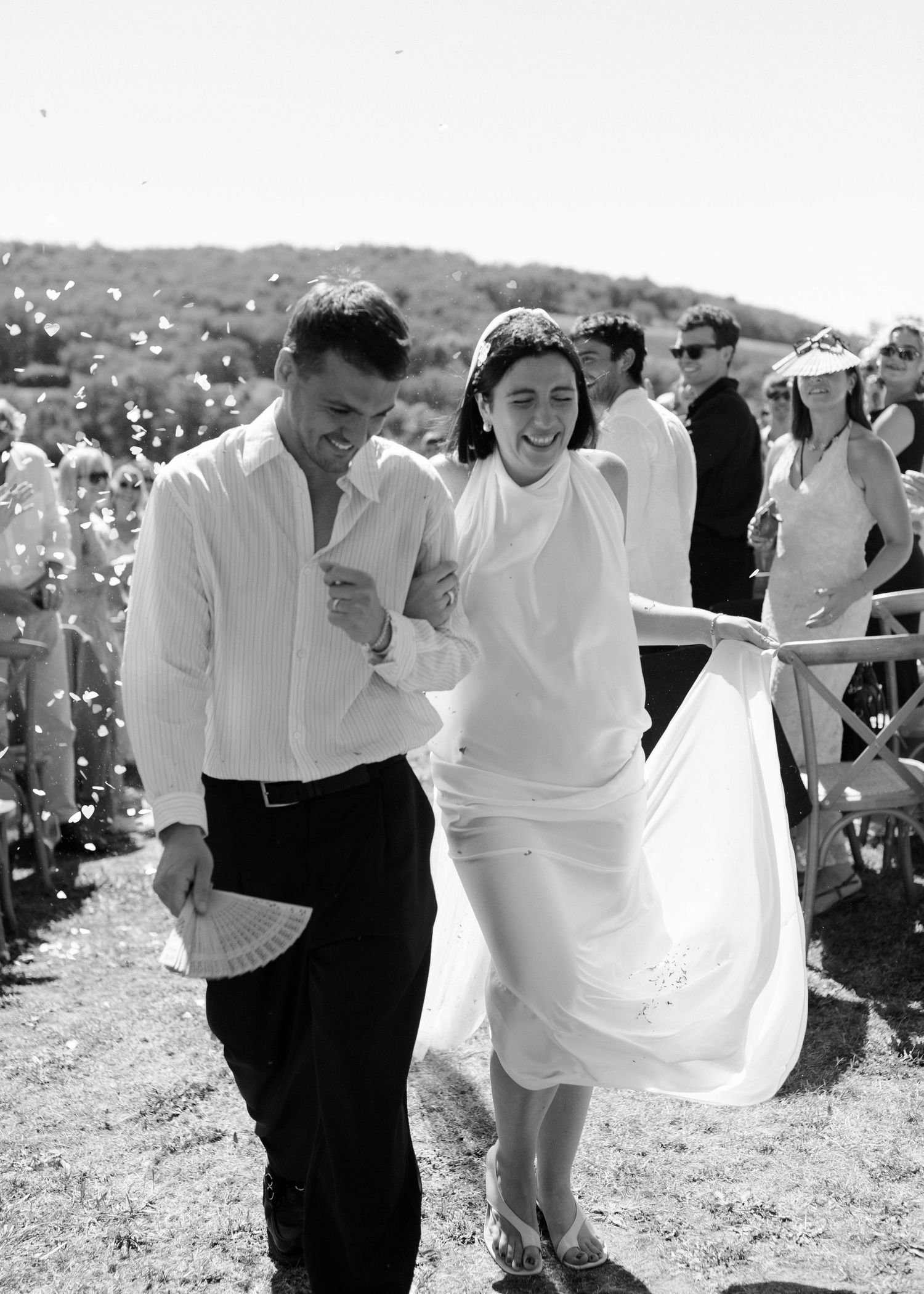A couple in wedding attire dances and twirls together in a sunny outdoor setting with guests in the background.