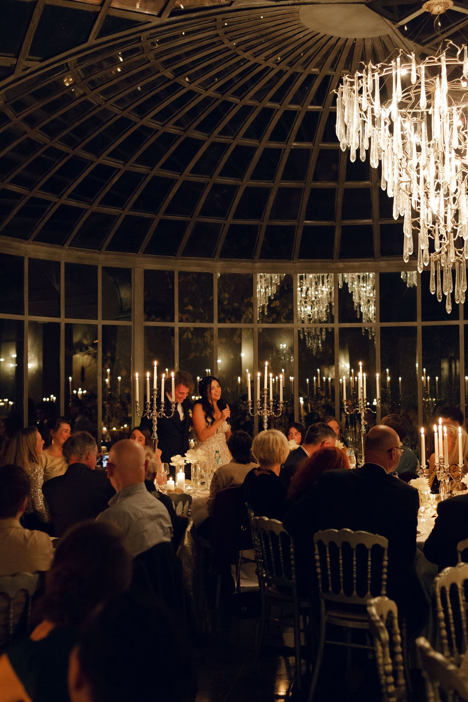 Elegant evening reception in a glass conservatory illuminated by crystal chandeliers and candlelight with guests seated at tables.